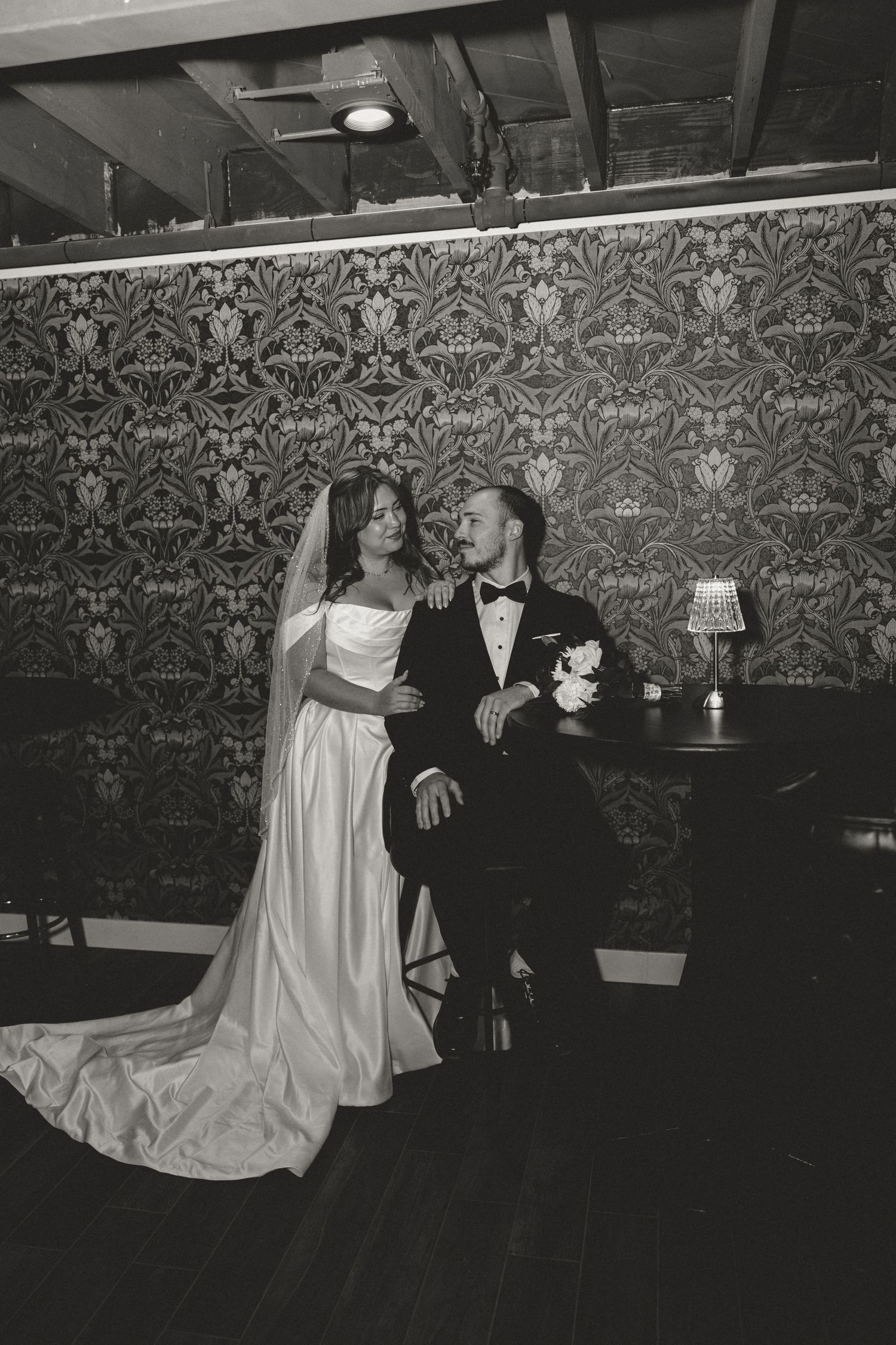 Black and white wedding portrait of bride in white gown and groom in tuxedo seated before ornate wallpaper.