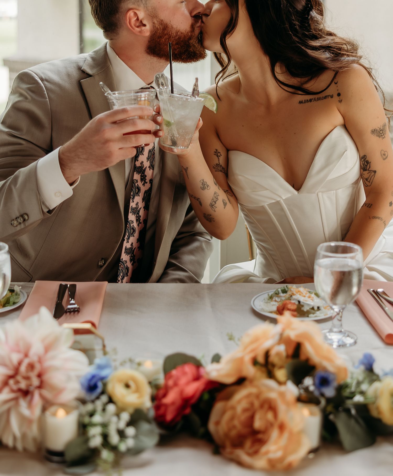 Bride and groom kissing while holding cocktails at wedding reception table with colorful floral centerpiece.