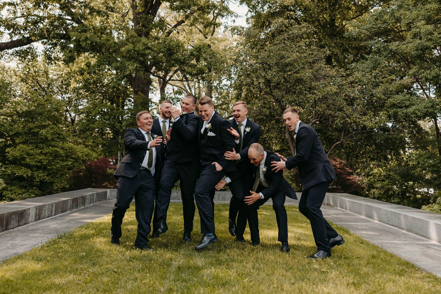 A group in matching black formal suits pose playfully together on stone steps surrounded by green trees in a garden setting.