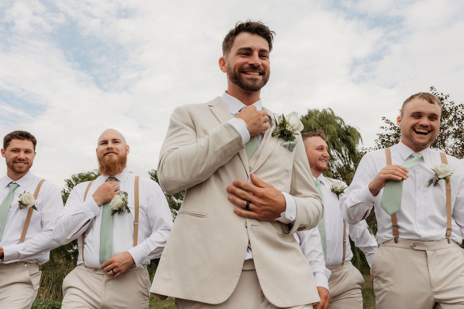 A group in matching beige and white suits laughs together at an outdoor wedding celebration.