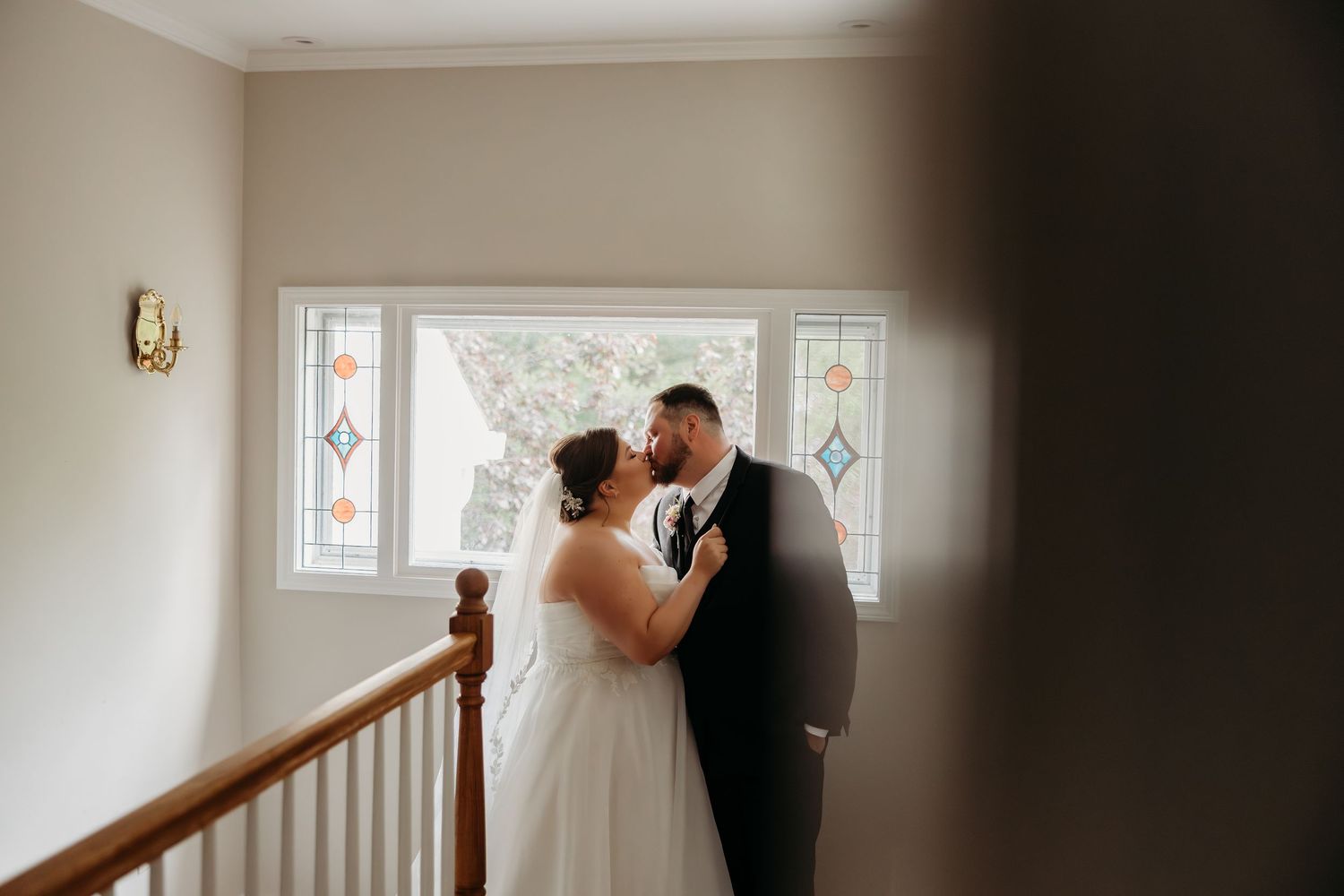 Bride and groom share a romantic first look kiss on a staircase landing with stained glass windows.