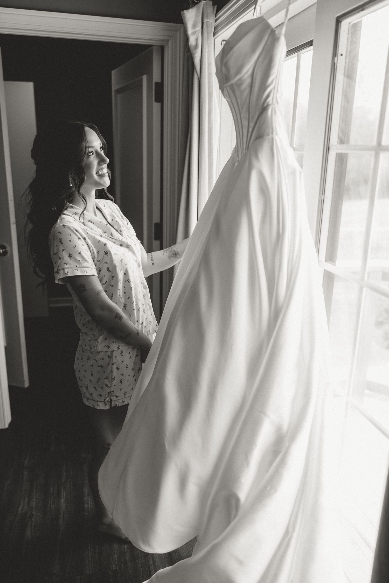 Black and white photo of a bride in a robe admiring her wedding gown hanging by a window.