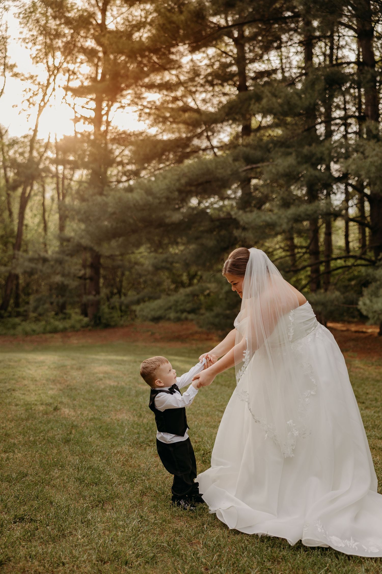 Bride in white gown and veil dancing with young ring bearer in black suit on green lawn at sunset.