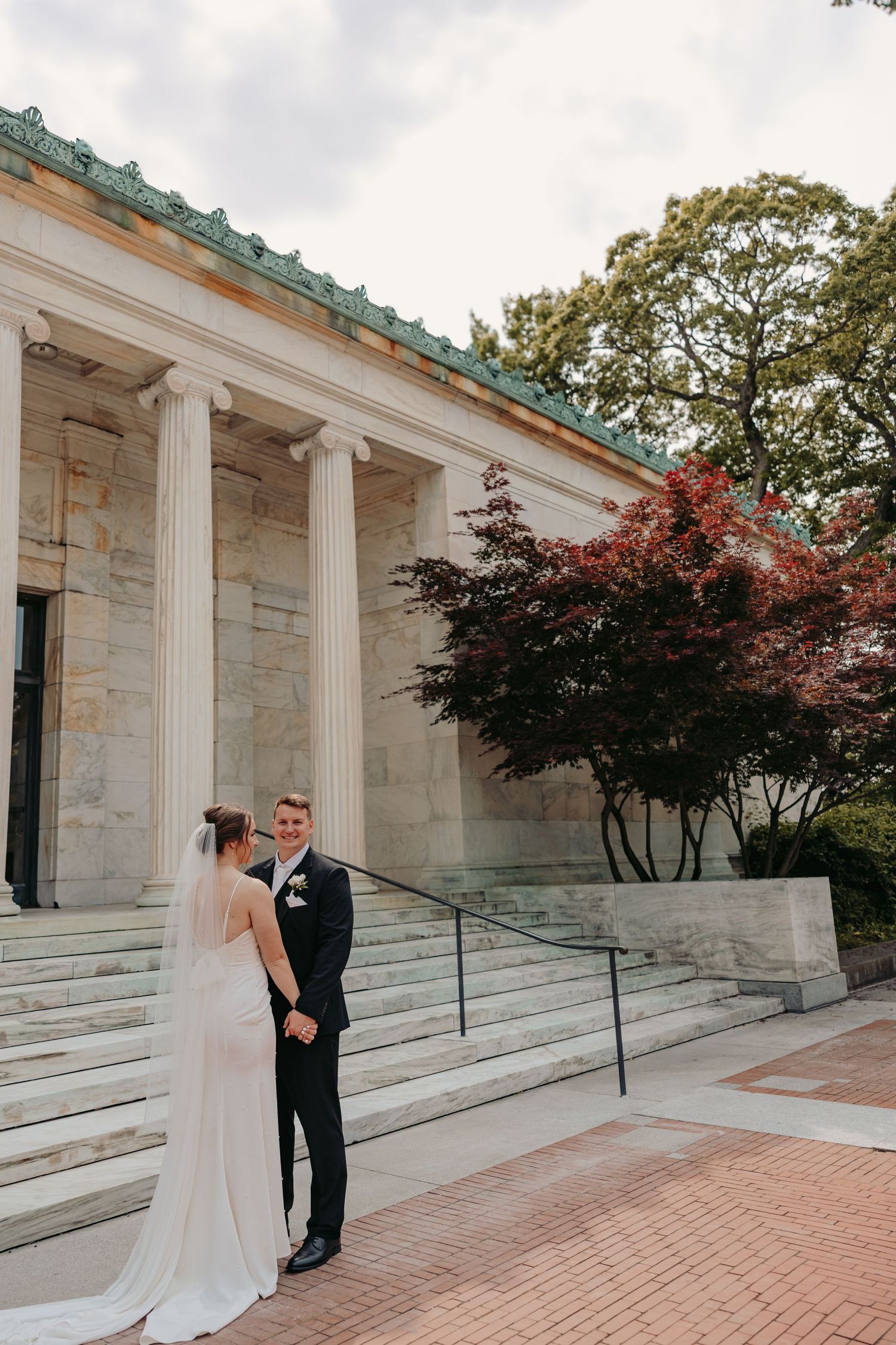 Wedding couple stands on brick steps in front of classical stone building with white columns and autumn foliage.