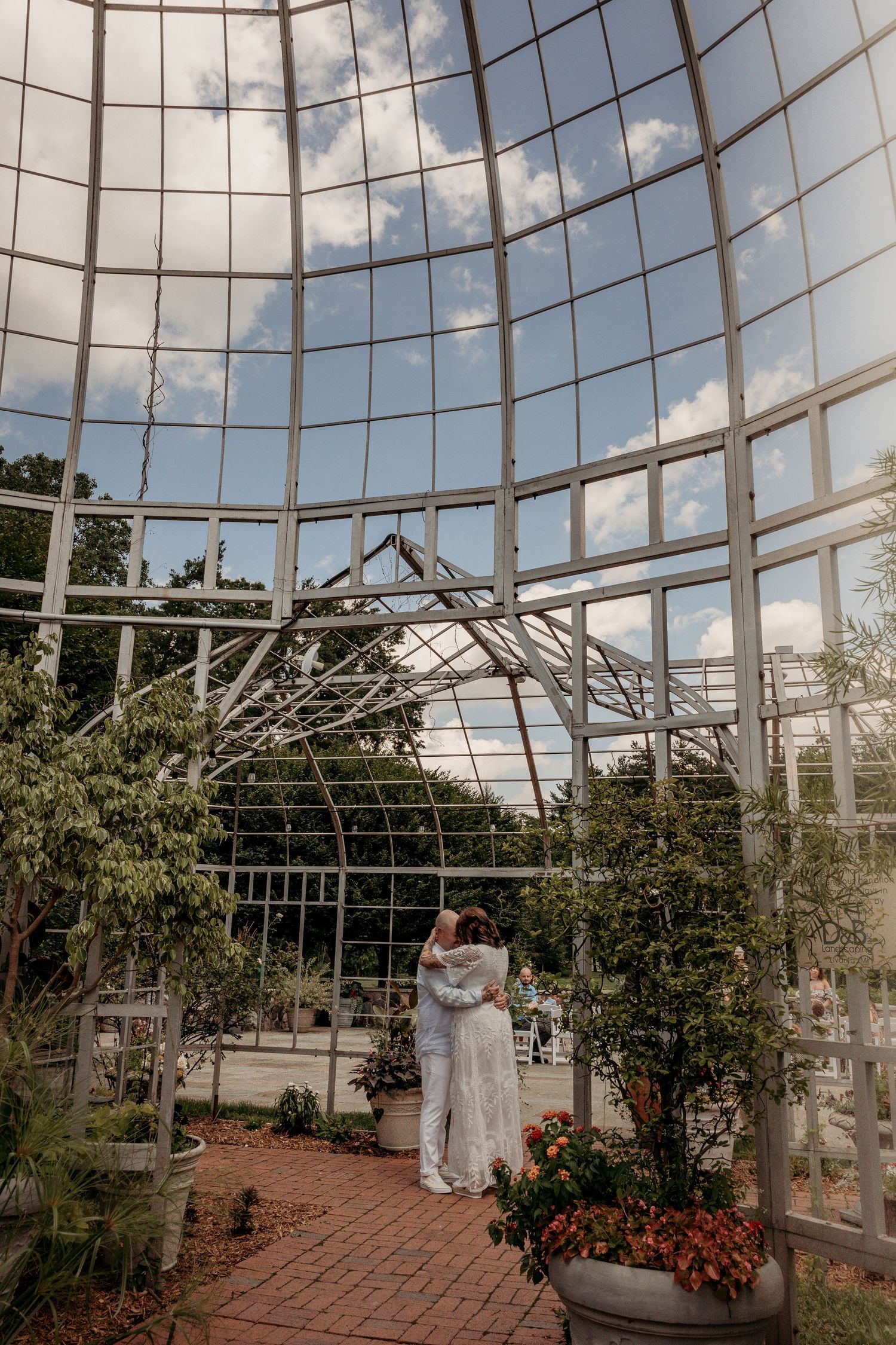 Ornate white greenhouse structure with curved glass panels and brick pathways surrounded by lush greenery against a cloudy sky.