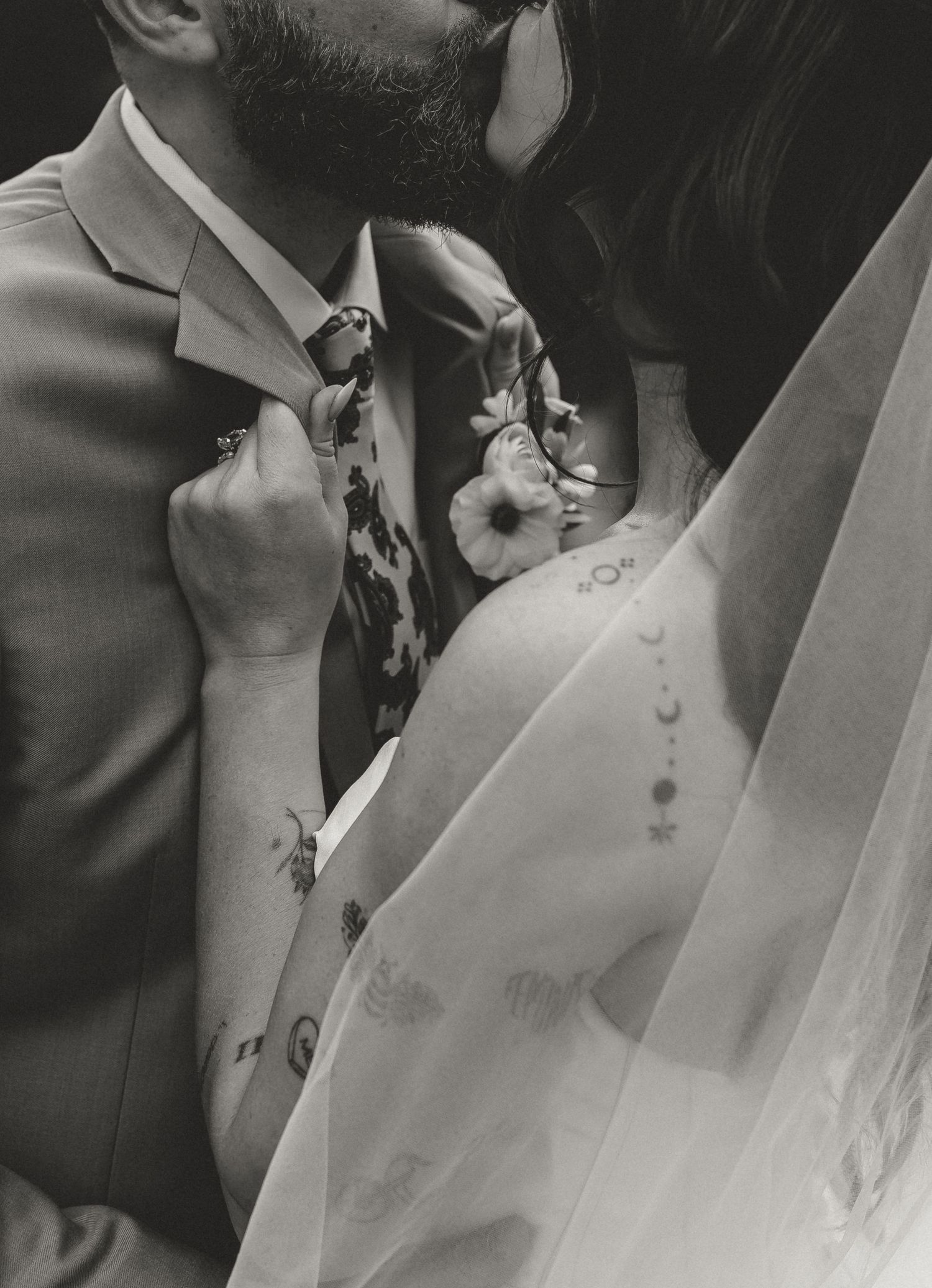 Black and white wedding photo of tattooed bride and groom sharing intimate moment, bride wearing veil with floral details.
