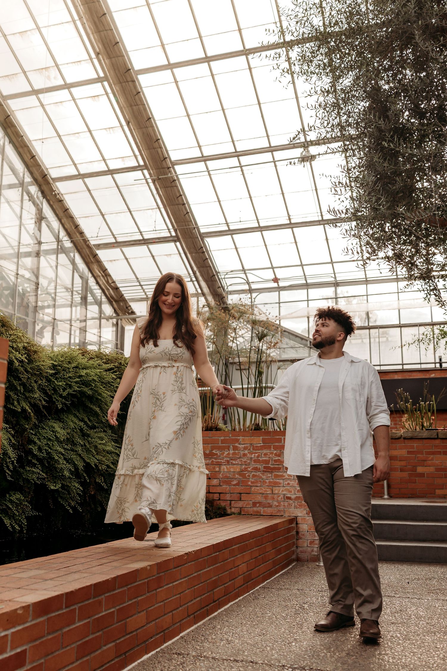 Two people walk together through a sunlit greenhouse with brick walls and gravel floors.