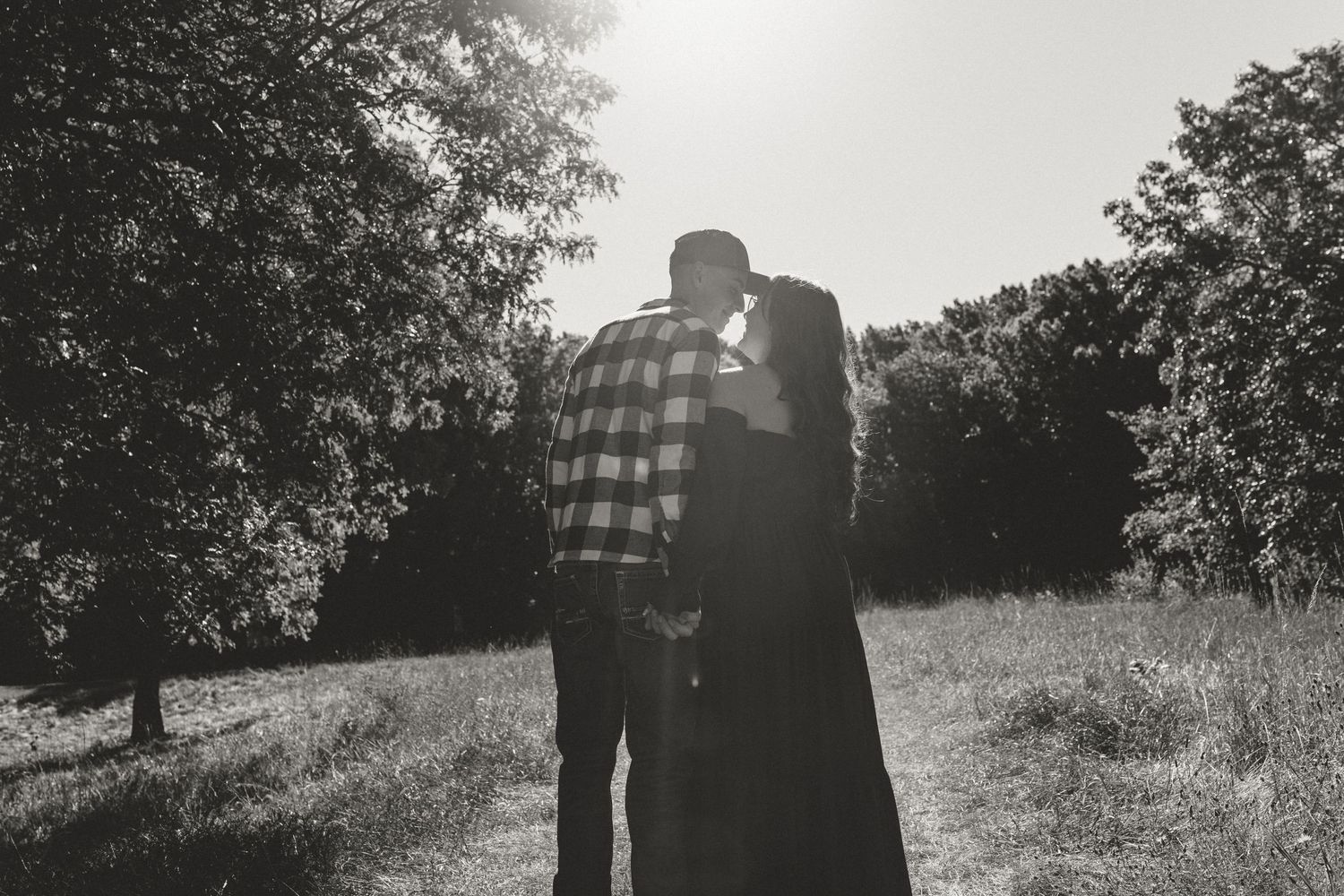 A couple in silhouette walks together through a sunny meadow with trees in the background.