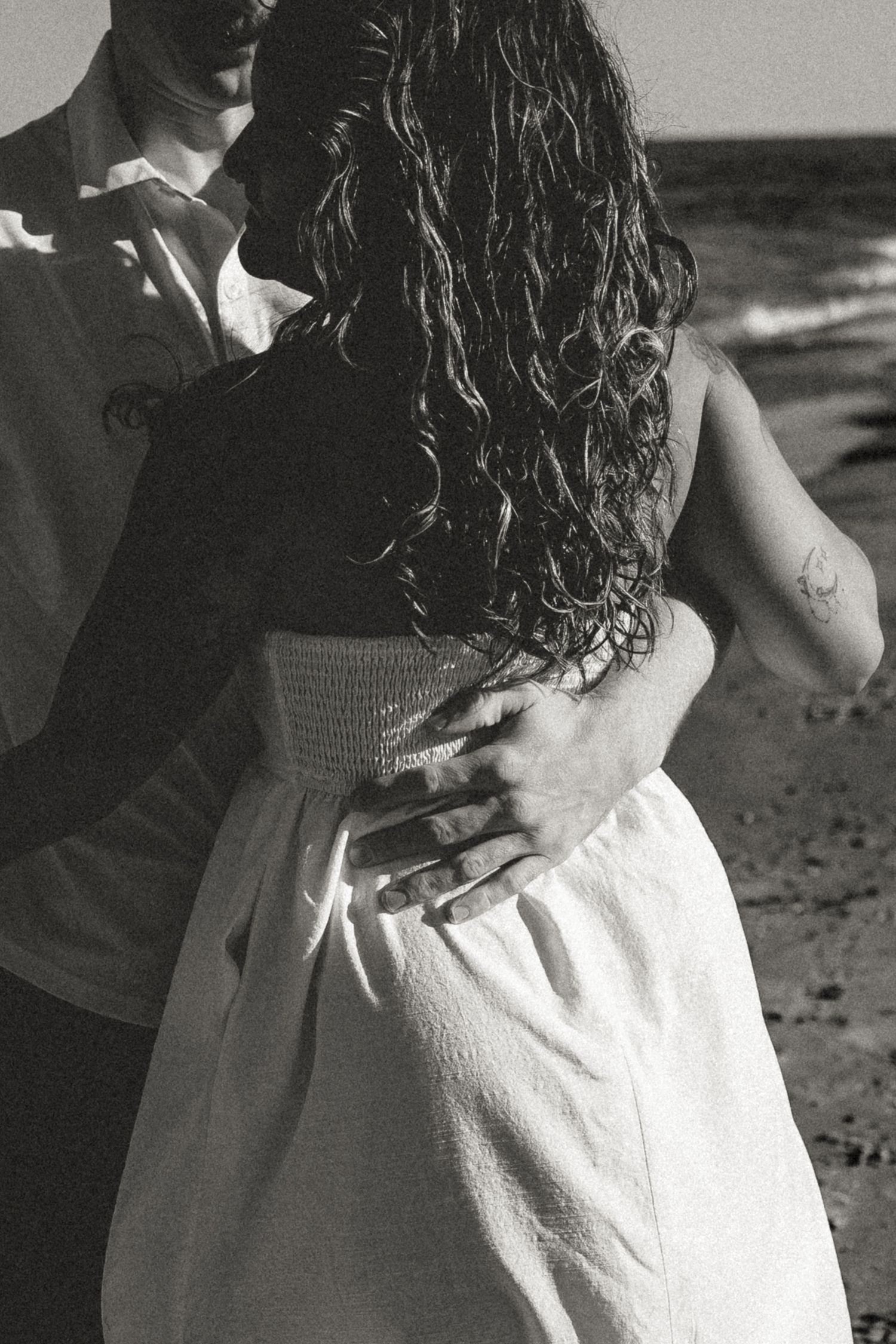A black and white photograph shows a person with curly hair wearing a light-colored dress standing on a beach.