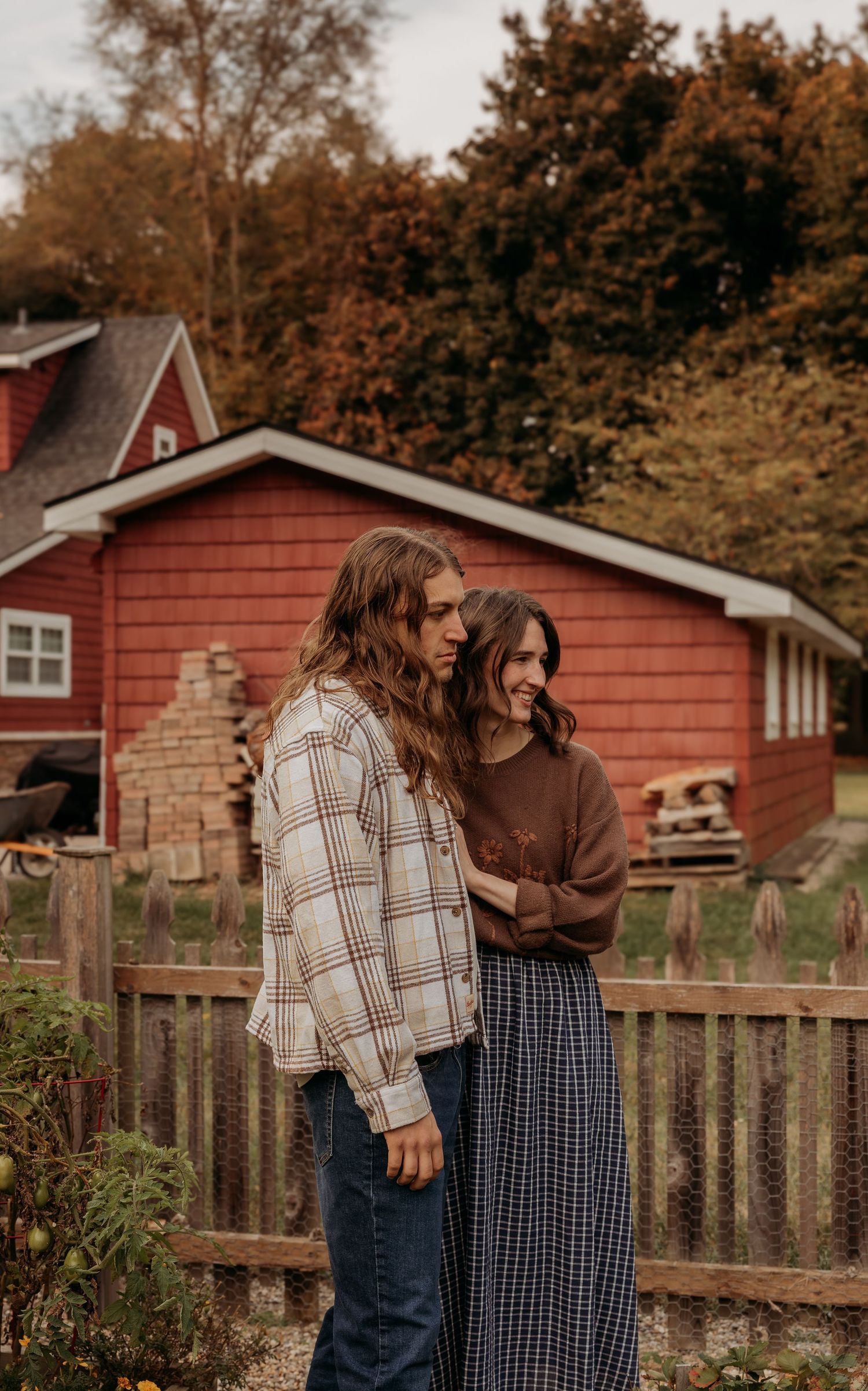 Two people share an intimate moment in front of red barn buildings on an autumn day.