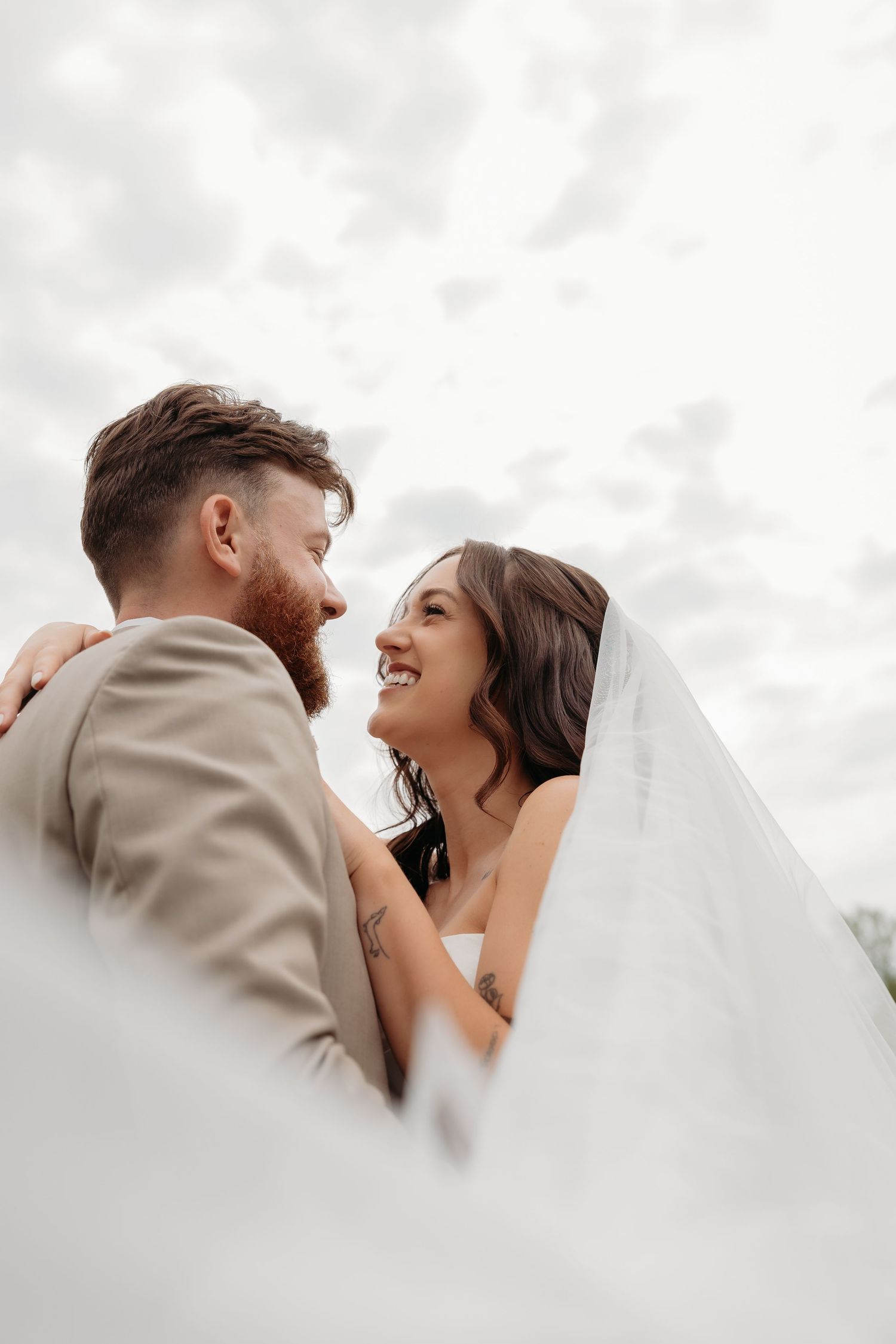Smiling bride and bearded groom in beige suit share romantic moment under cloudy sky on wedding day.