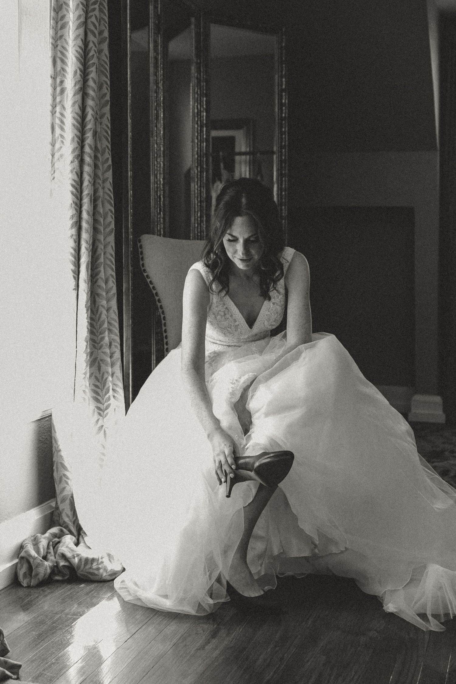 A black and white photograph shows a figure in a flowing white wedding gown adjusting their shoes in an elegant room.