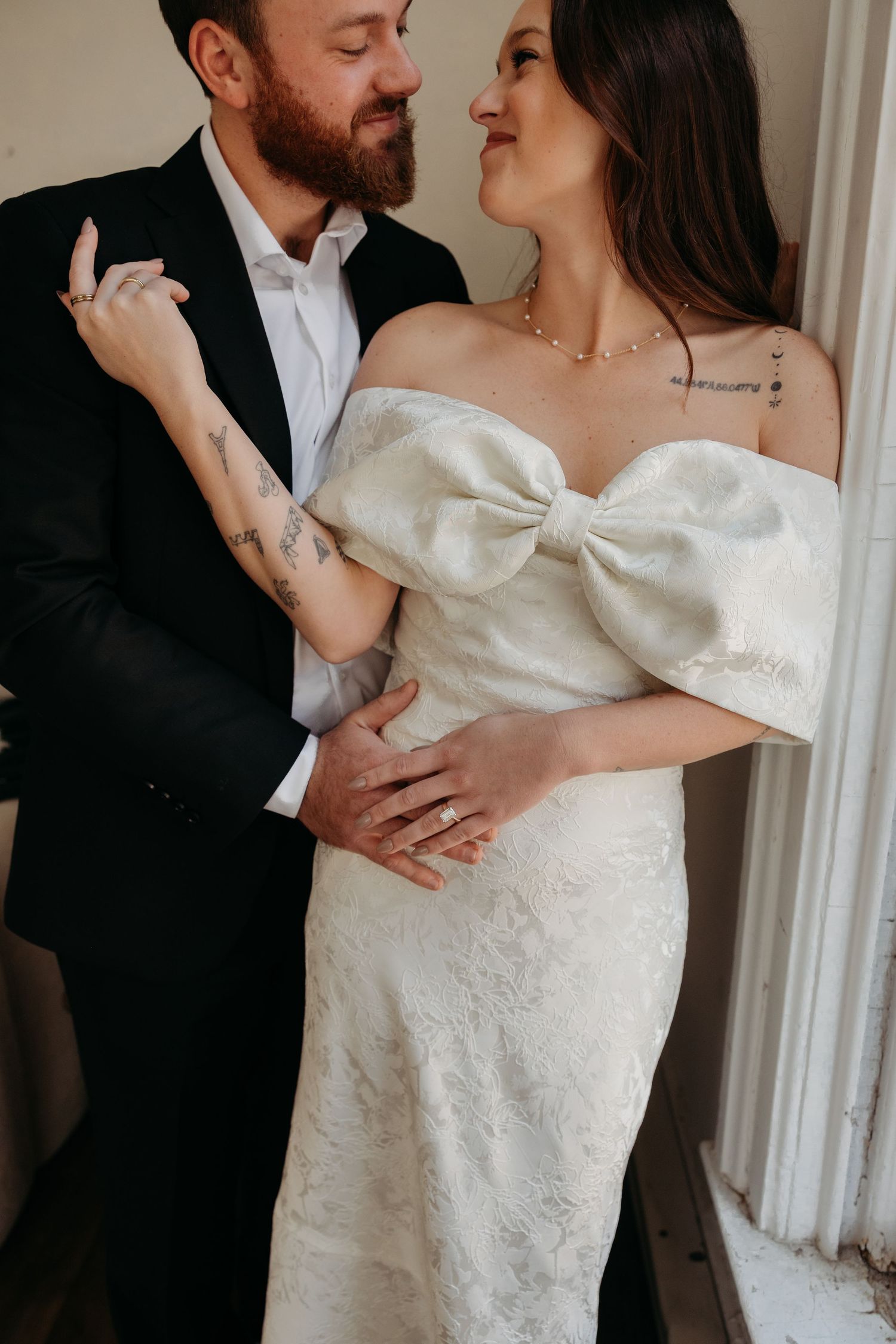 Intimate wedding portrait showing couple embracing by window, with bride in white off-shoulder gown with bow detail.