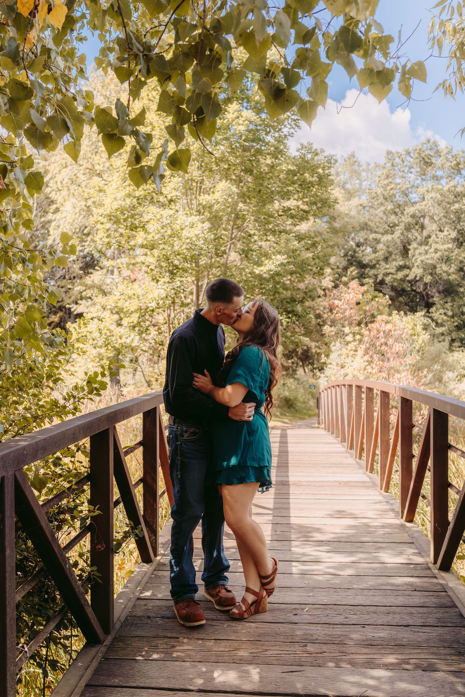 A romantic couple shares an intimate moment on a wooden boardwalk surrounded by lush summer foliage.