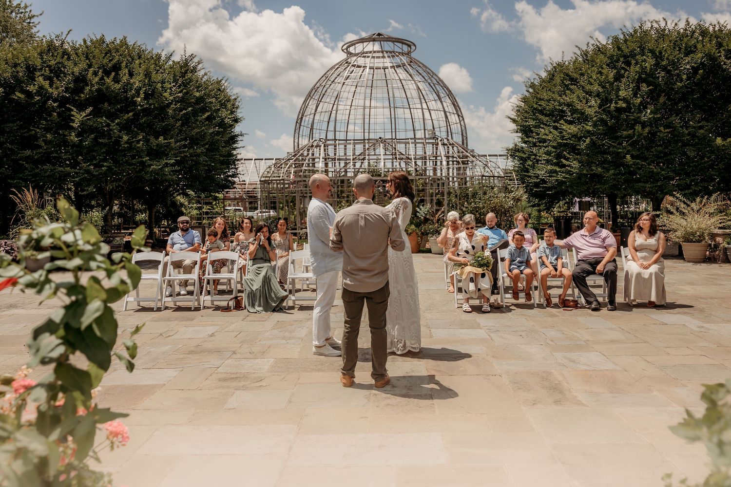 An outdoor wedding ceremony takes place in front of a grand glass conservatory dome with guests seated on white chairs.