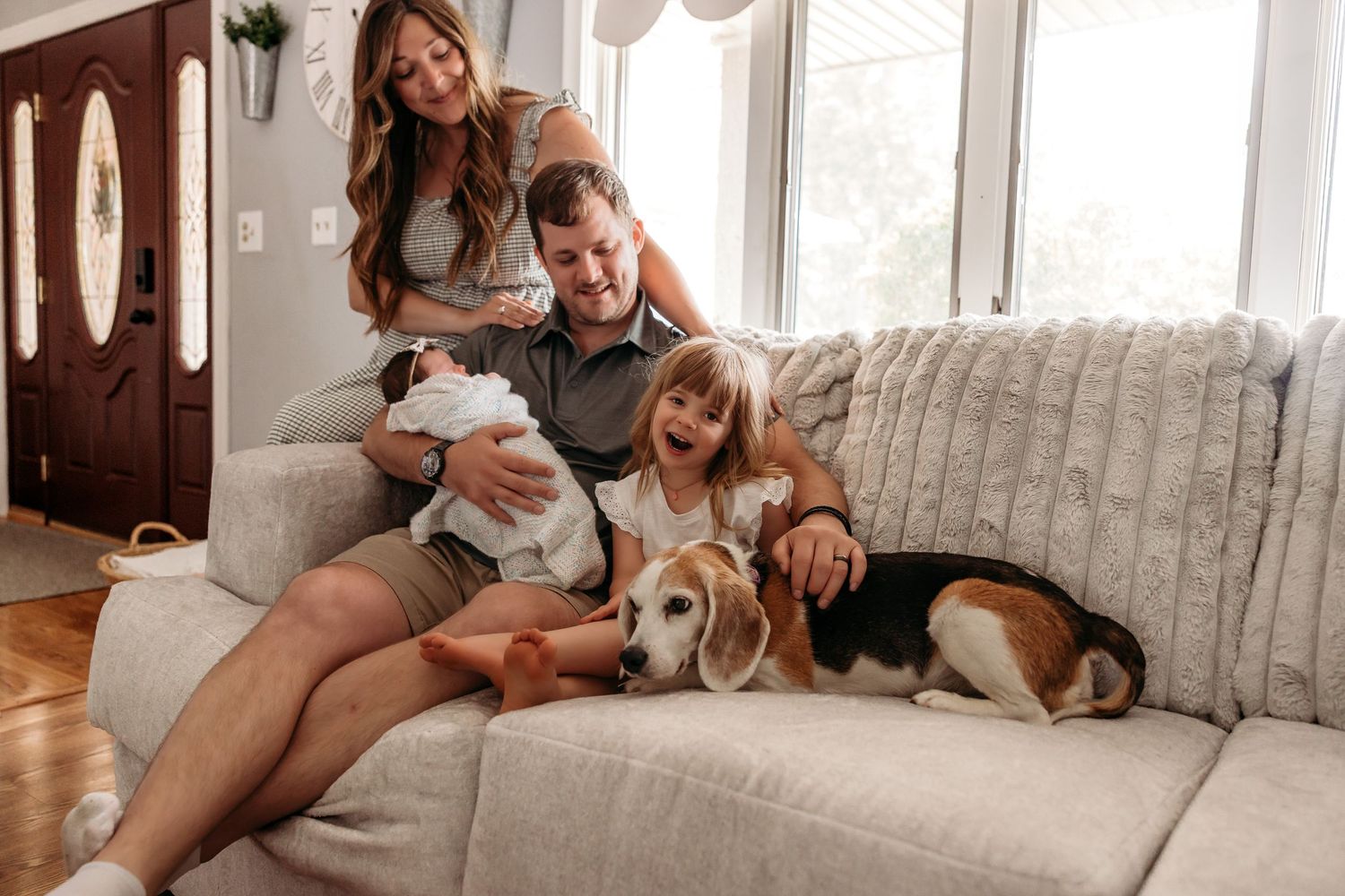 A family lounges together on a light gray couch with their beagle dog in a sunny living room.