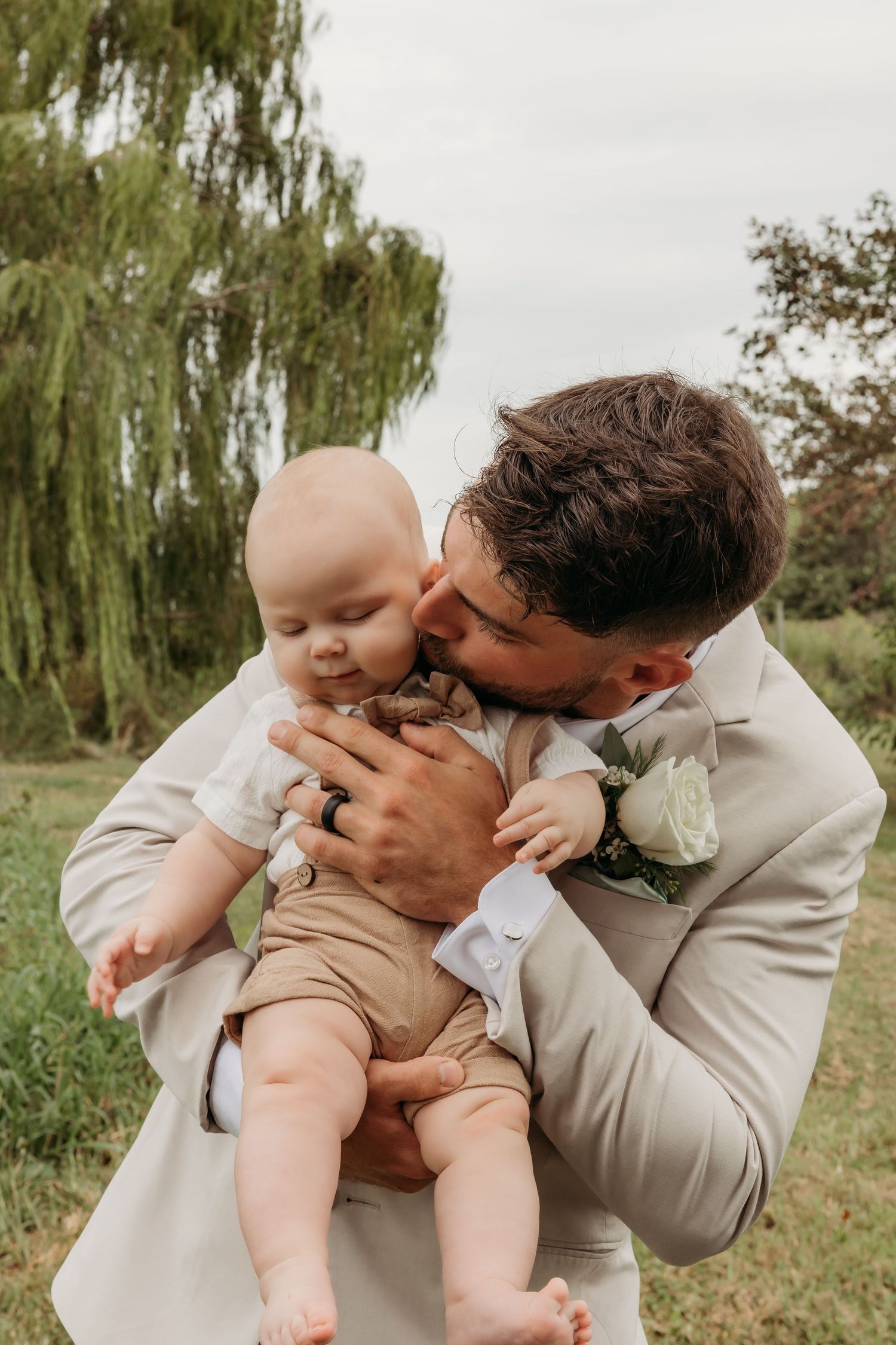 A parent in beige suit holding and kissing their baby in a green outdoor setting under a willow tree.