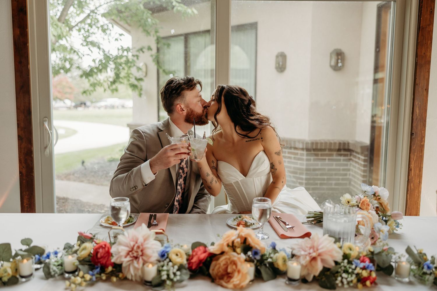 Bride and groom share a romantic kiss at floral-decorated sweetheart table during indoor wedding reception.