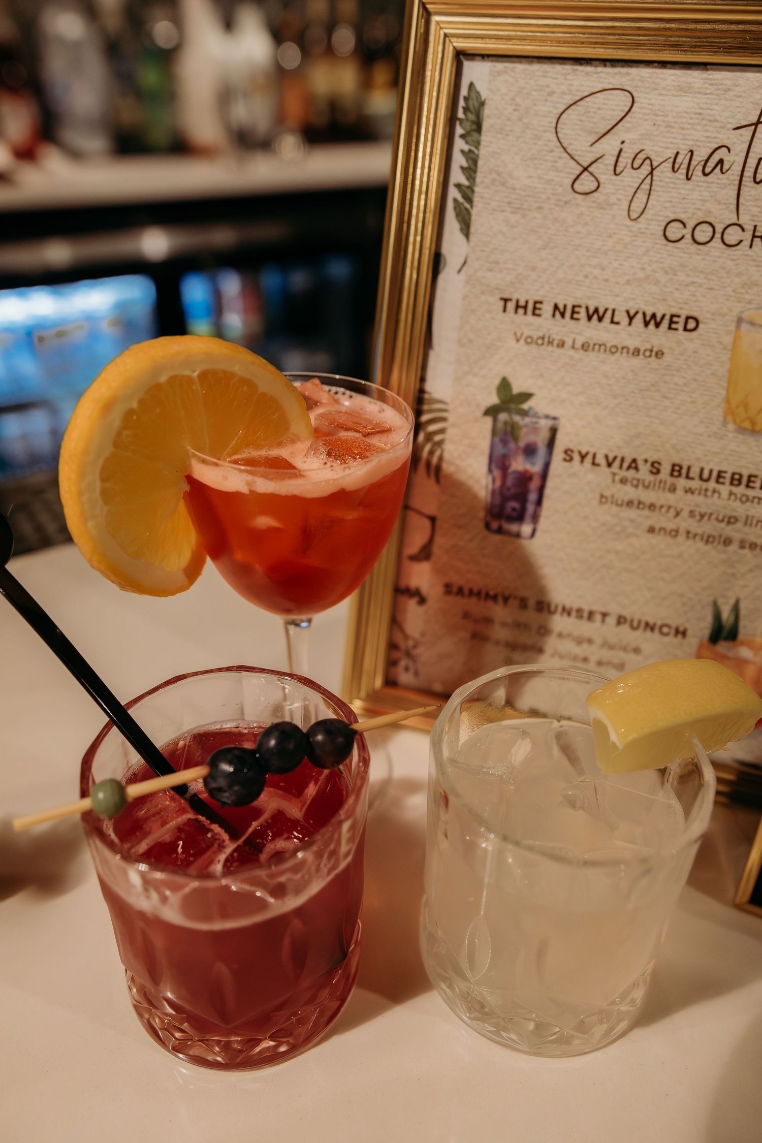 Signature cocktail menu display with three drinks including a red cocktail, blueberry drink, and lemonade at a wedding bar.