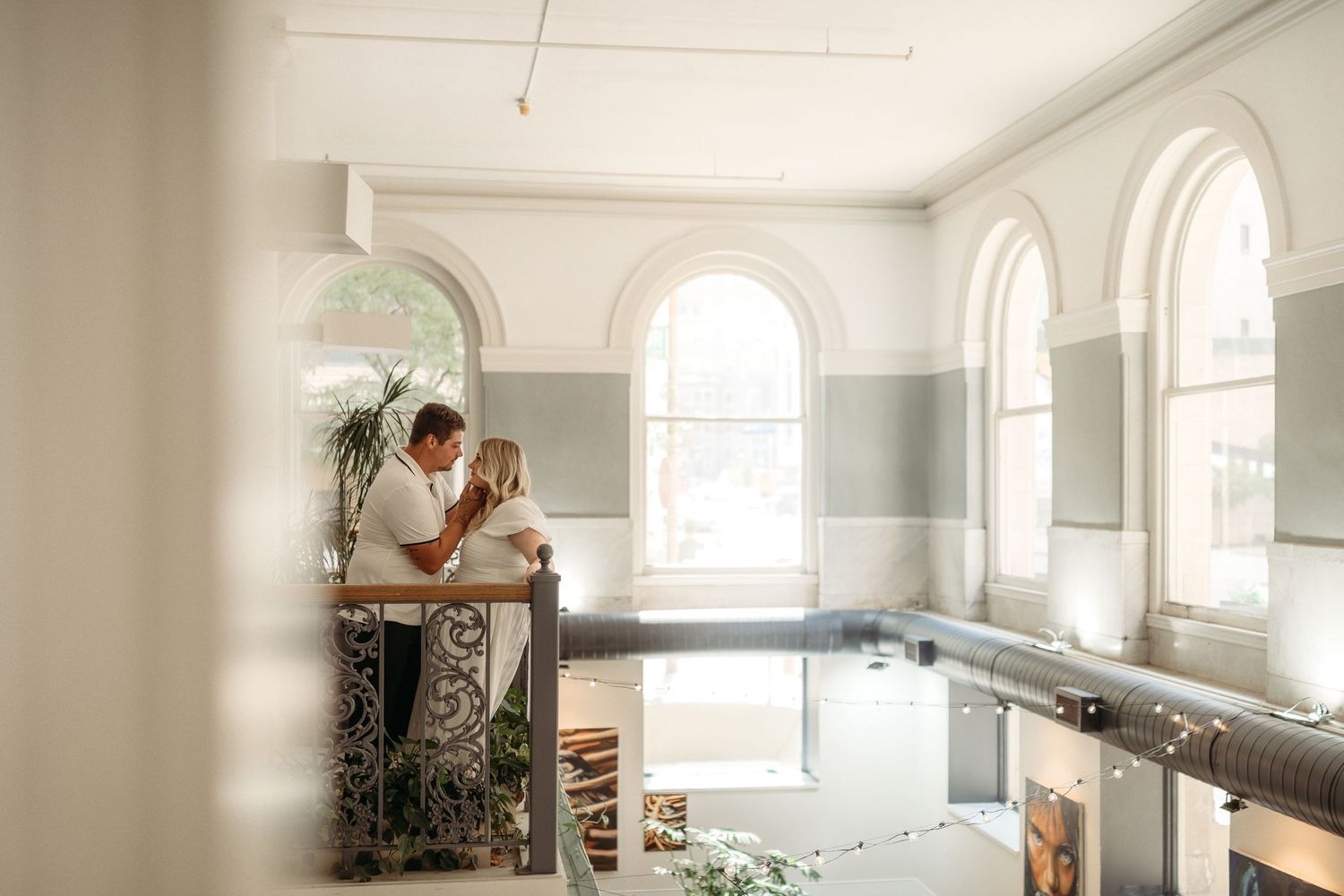Two people stand on an elegant staircase landing with arched windows and ornate railings in a bright interior space.