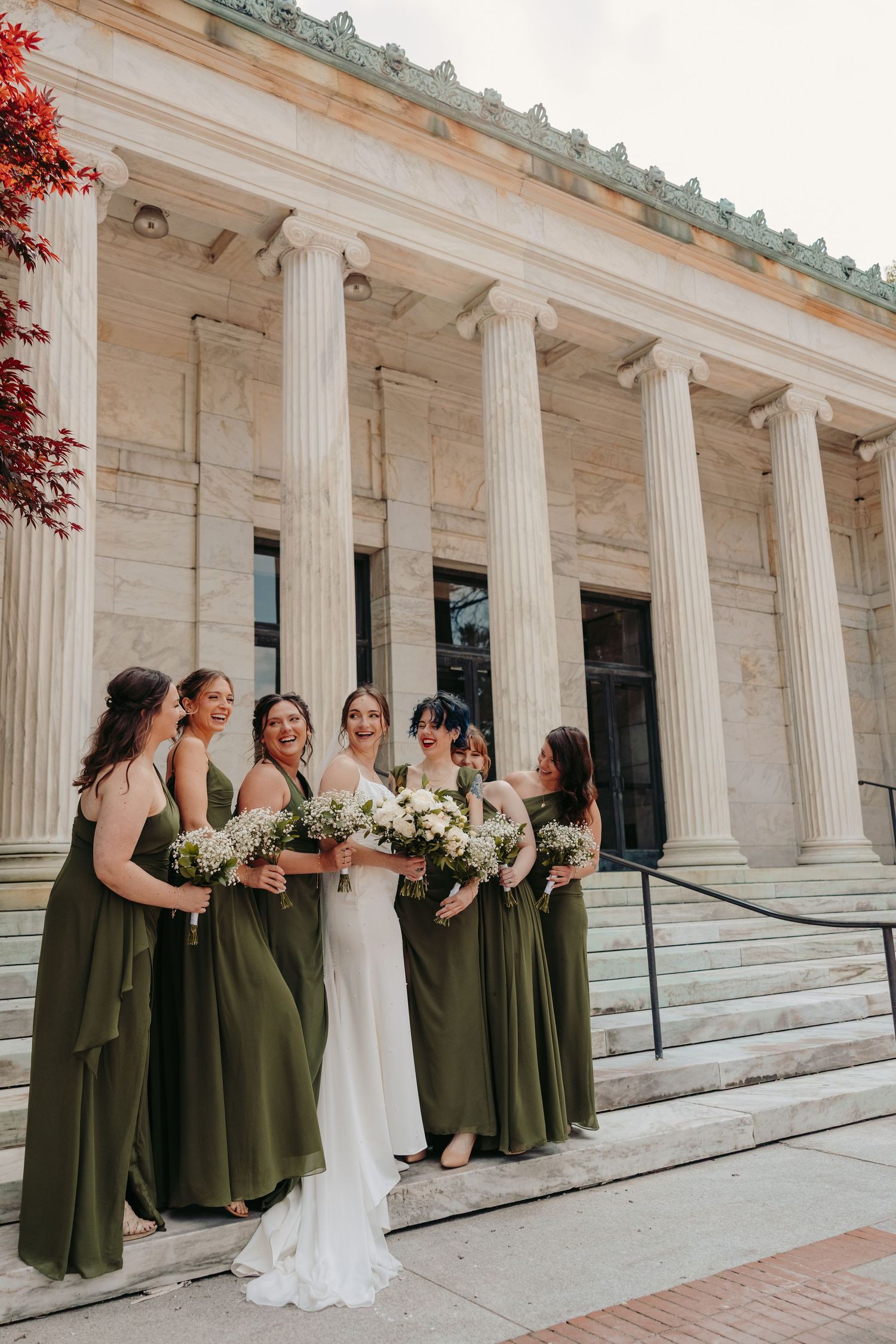 A wedding party in olive green bridesmaid dresses posed along white marble columns of a classical building.