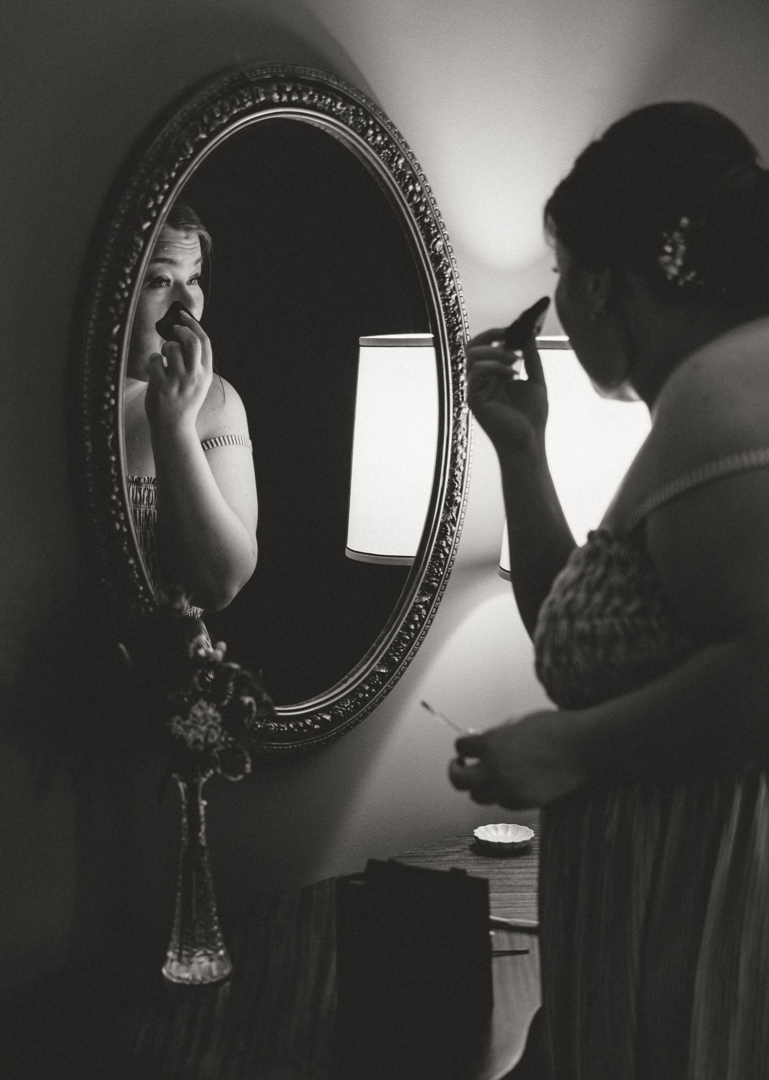 Black and white photo of a woman applying makeup while looking into an ornate oval mirror in a dimly lit room.
