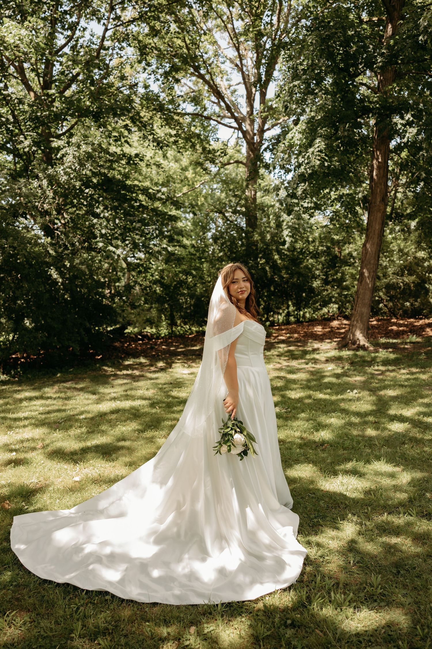 Bride in white off-shoulder gown with veil and bouquet standing on green lawn surrounded by lush trees.