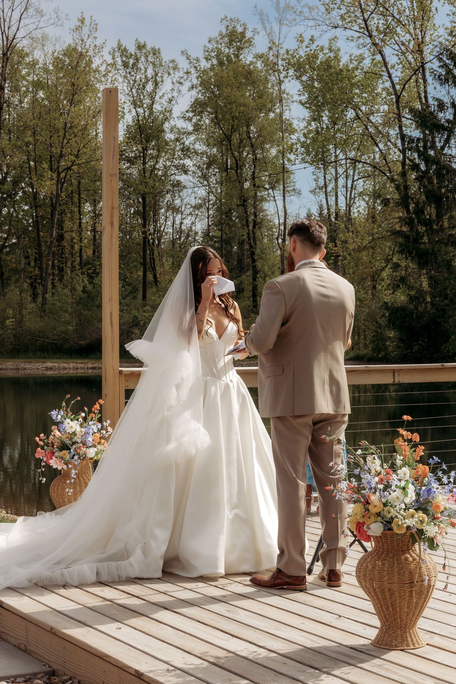 Bride and groom exchange vows on sunlit dock overlooking lake, surrounded by colorful floral arrangements.