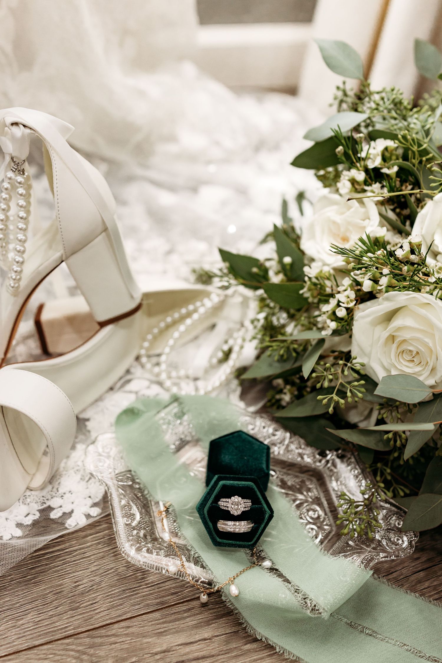 Wedding rings nestled in a velvet green ring box surrounded by white roses and greenery on a rustic wooden surface.