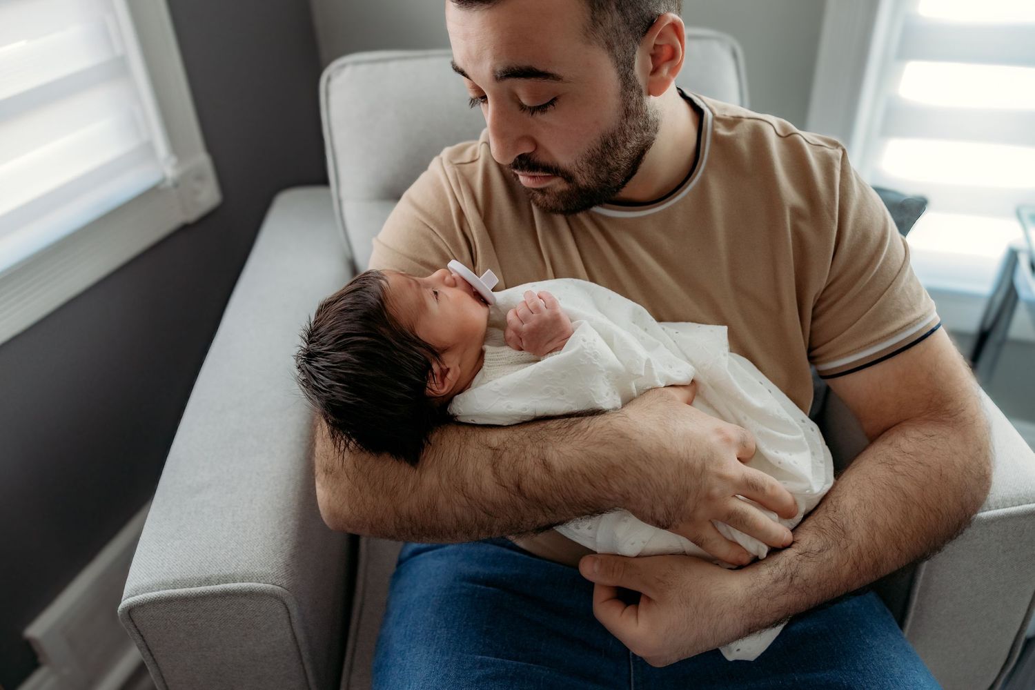 Adult holding and cradling sleeping newborn baby in casual clothing while sitting by window.