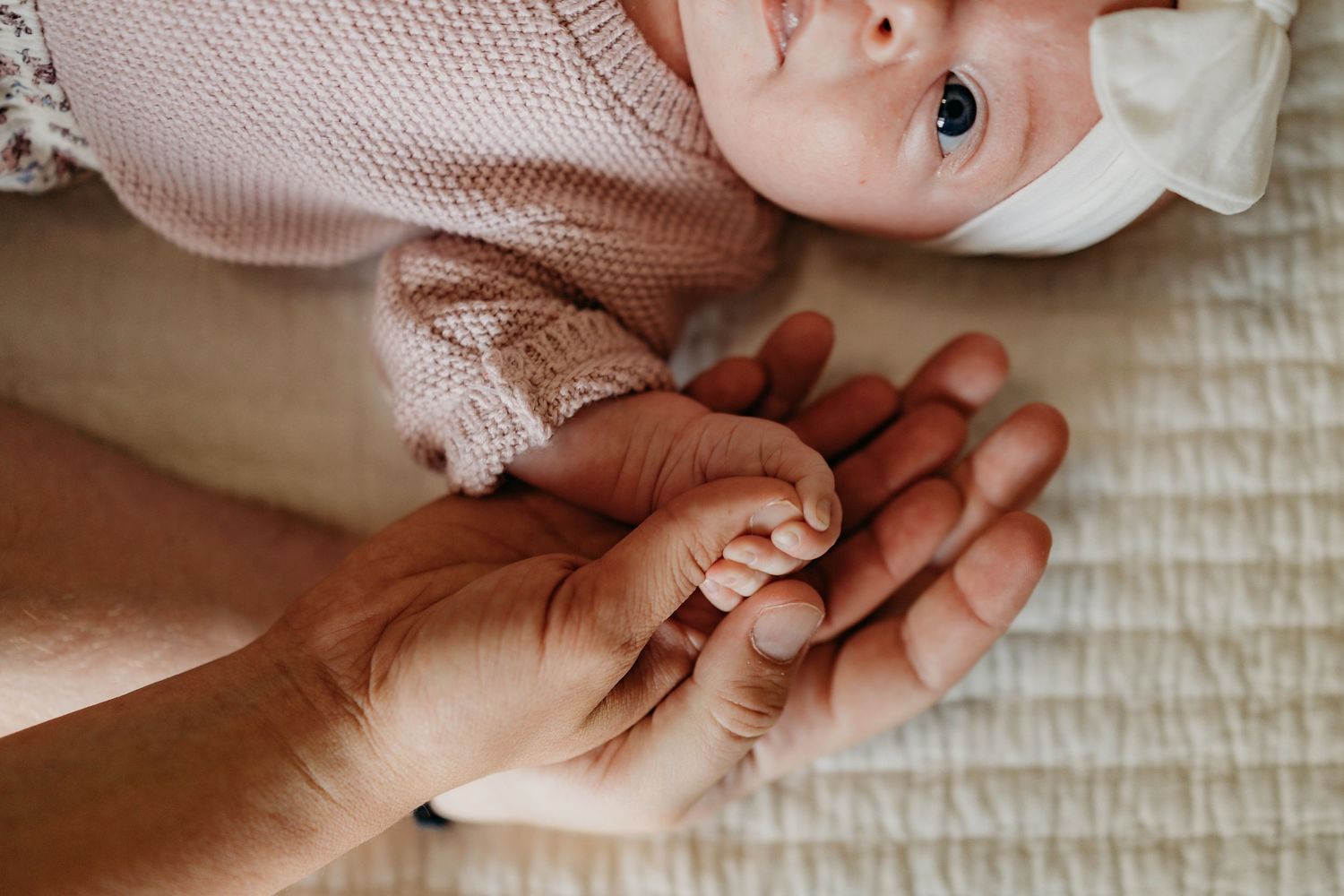 A close-up tender moment shows new parents holding their six-month-old baby's hand against a neutral background.