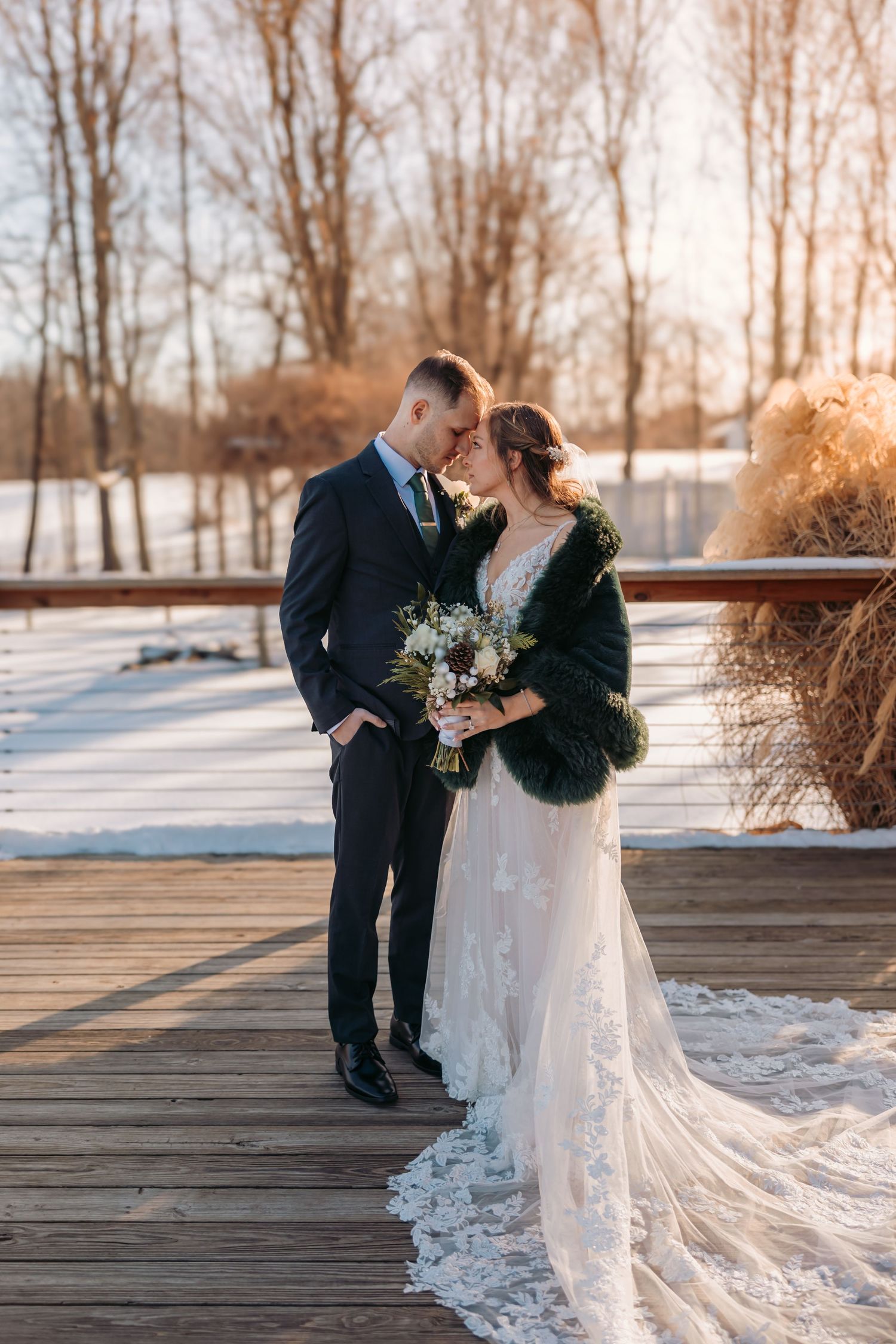 A romantic winter wedding couple embraces on a wooden deck surrounded by snow and bare trees at sunset.