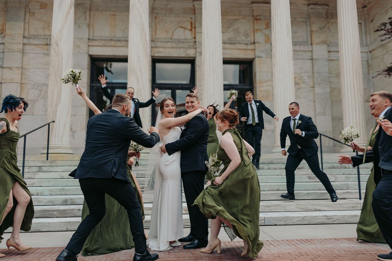 Wedding party members perform dramatic dance poses on grand building steps with white columns.