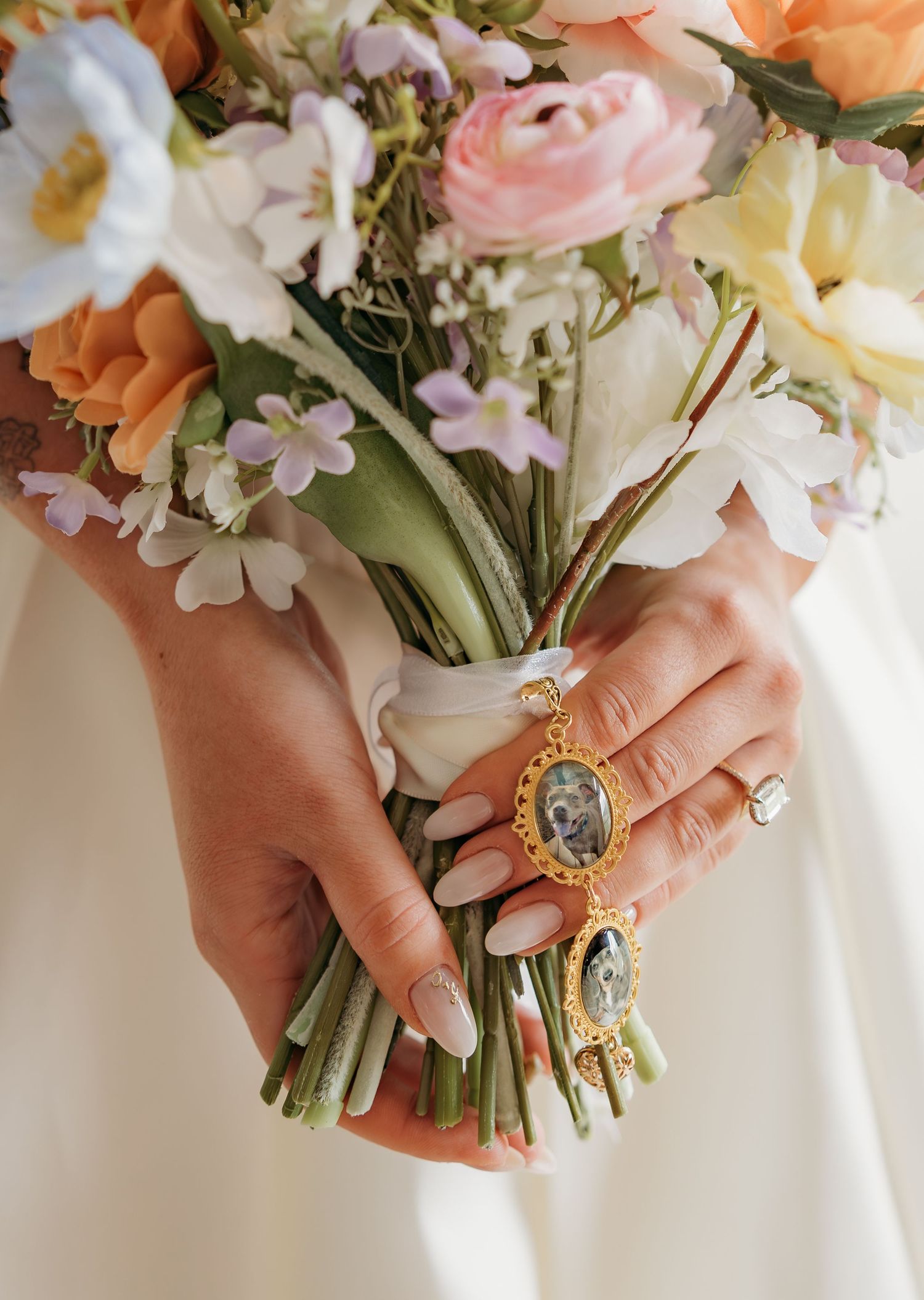 Bride holding a colorful wedding bouquet with a gold locket charm, wearing a diamond ring and white gown.