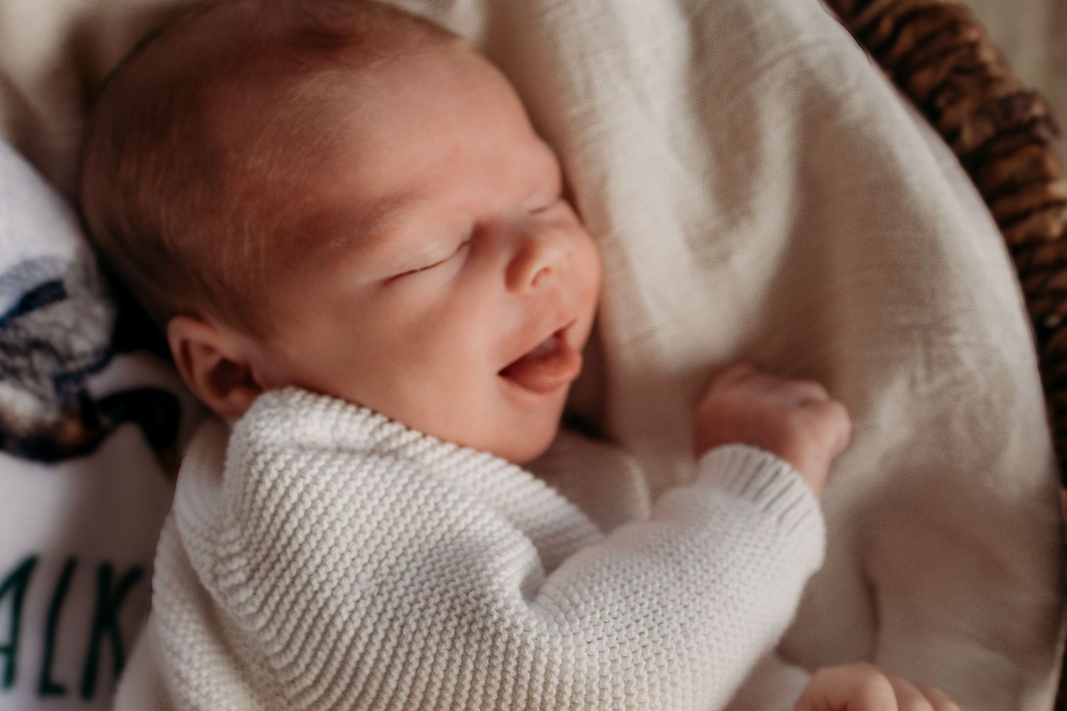 A sleeping newborn baby wrapped in a cozy white knit sweater smiles while resting in a woven basket.