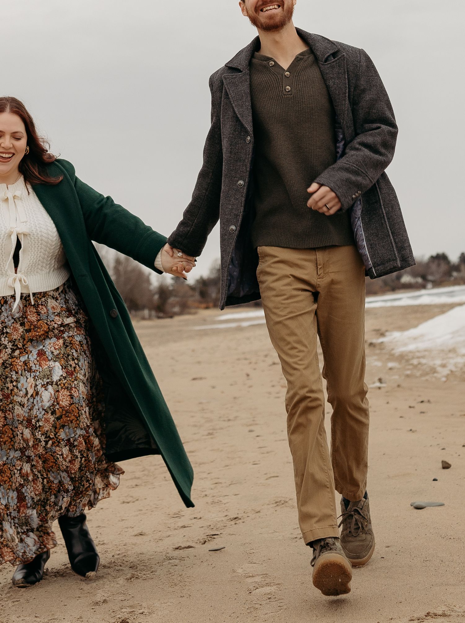 Couple walking and laughing together on a sandy beach wearing casual winter attire.