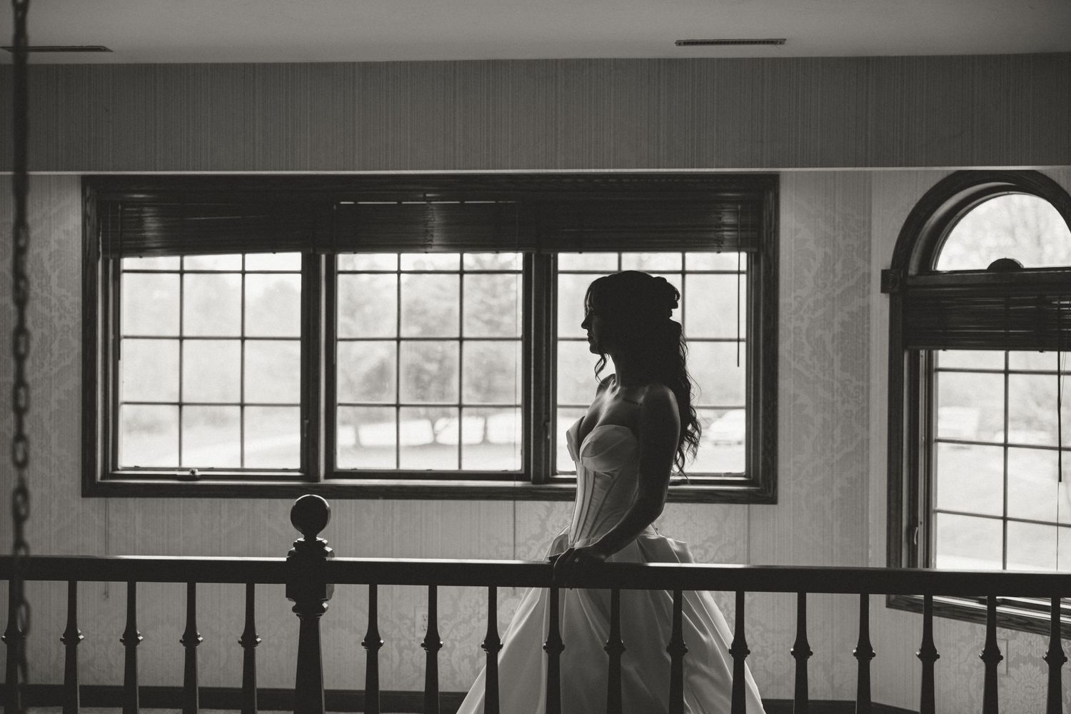 Bride in wedding dress stands by large windows on balcony in elegant black and white portrait photo.