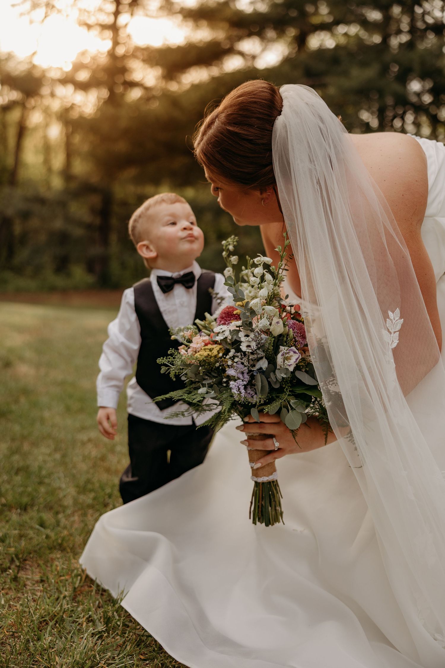 Bride in white gown and veil holds floral bouquet while sharing a sweet moment with a young boy in a bow tie outdoors.