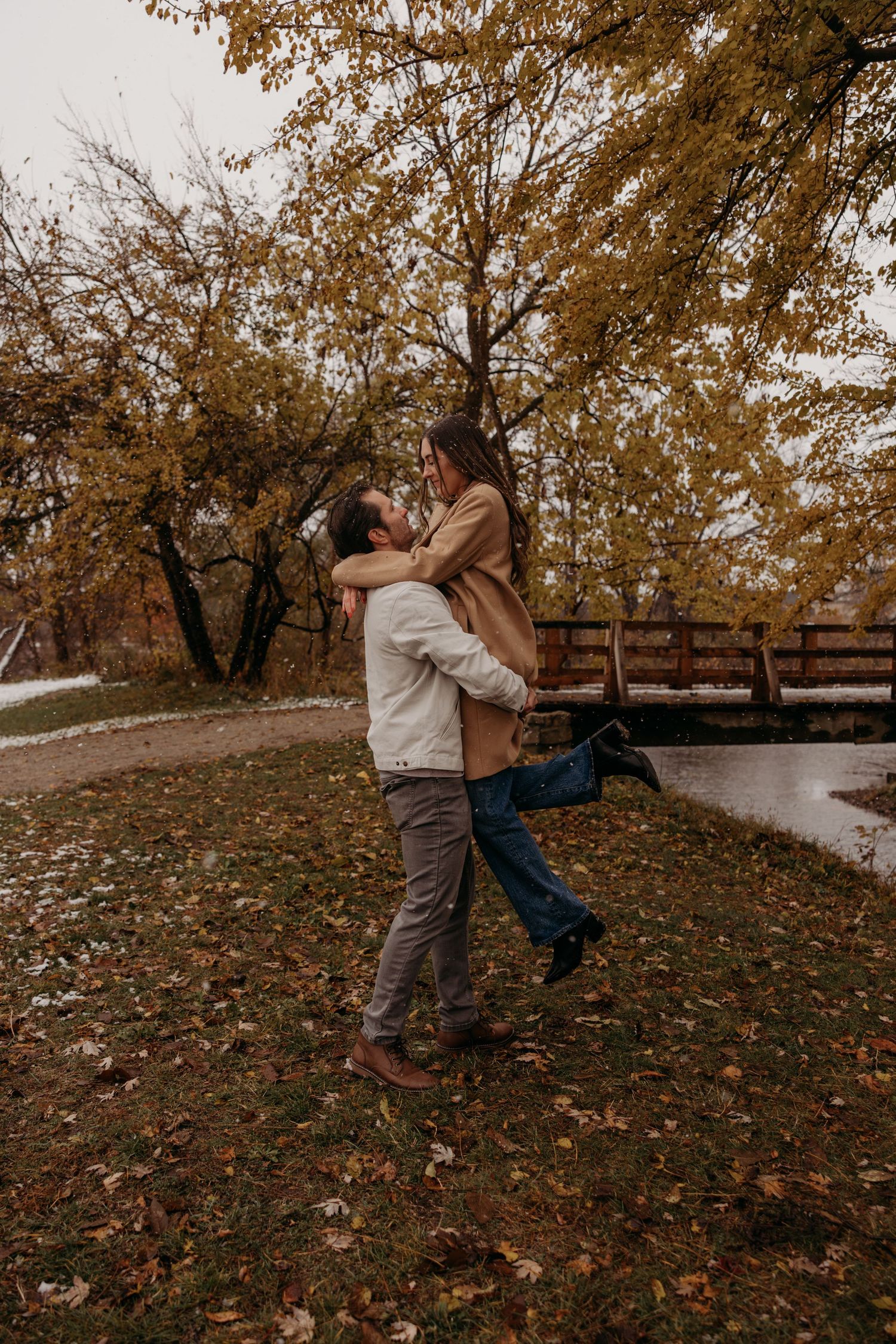 A playful autumn engagement photoshoot with a couple swinging in a leaf-covered park setting.