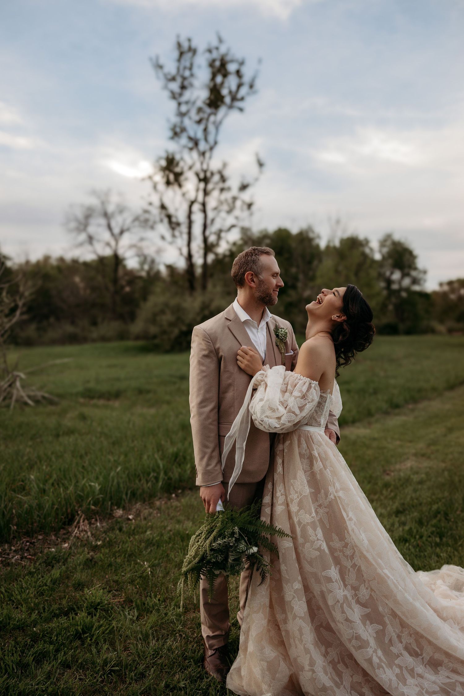 A couple in wedding attire laugh together in a lush green field at sunset while holding a rustic bouquet.