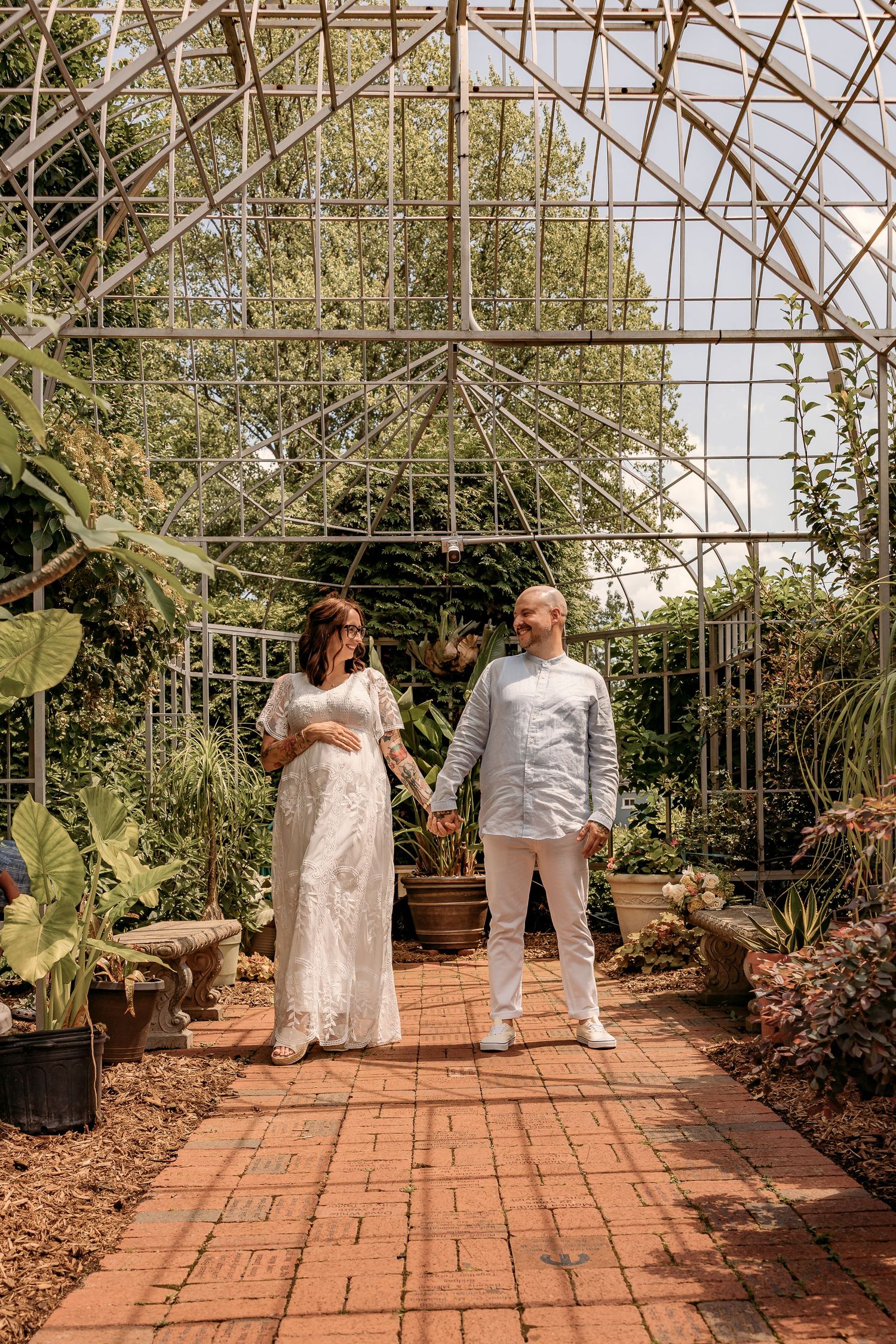Couple stands on brick path in a sunlit greenhouse filled with lush plants and greenery.