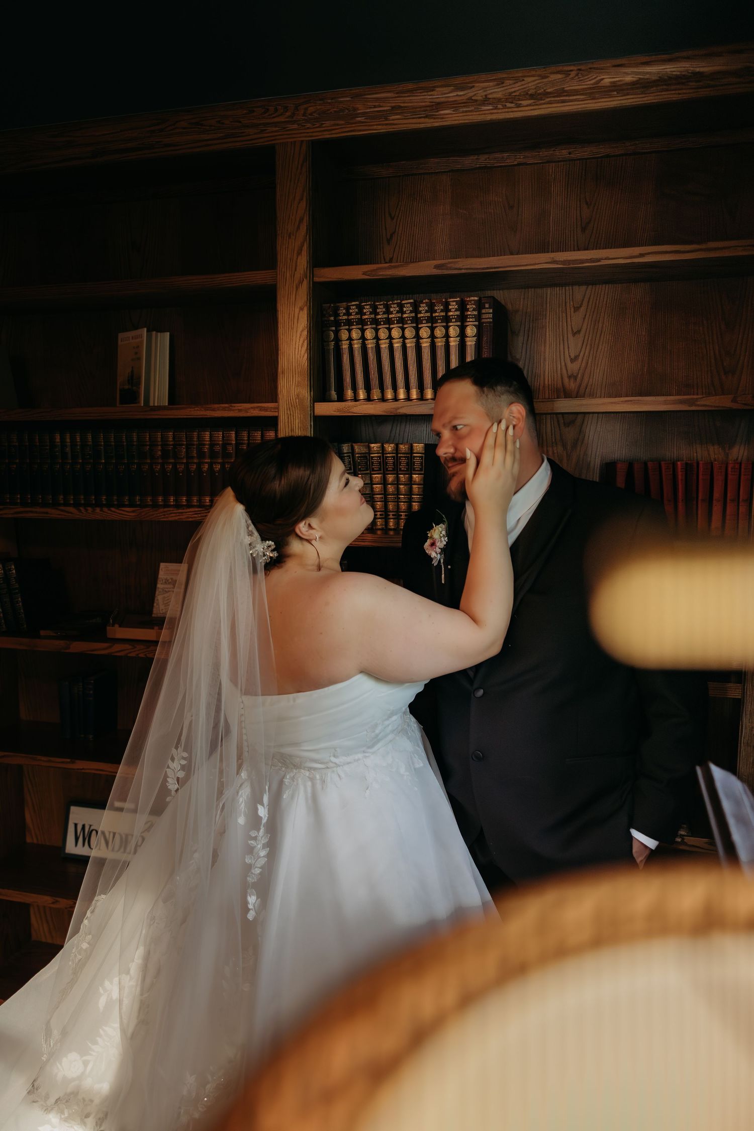 Bride in white gown and veil tenderly touches groom's face in romantic library setting with wooden bookshelves.