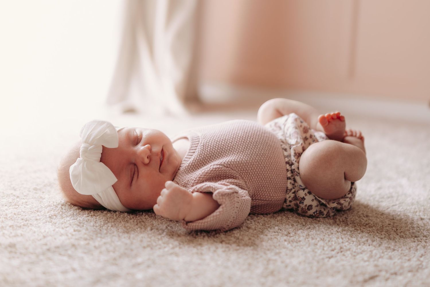 Baby wearing cozy knit outfit and bow headband rests peacefully on beige carpet in soft natural lighting.