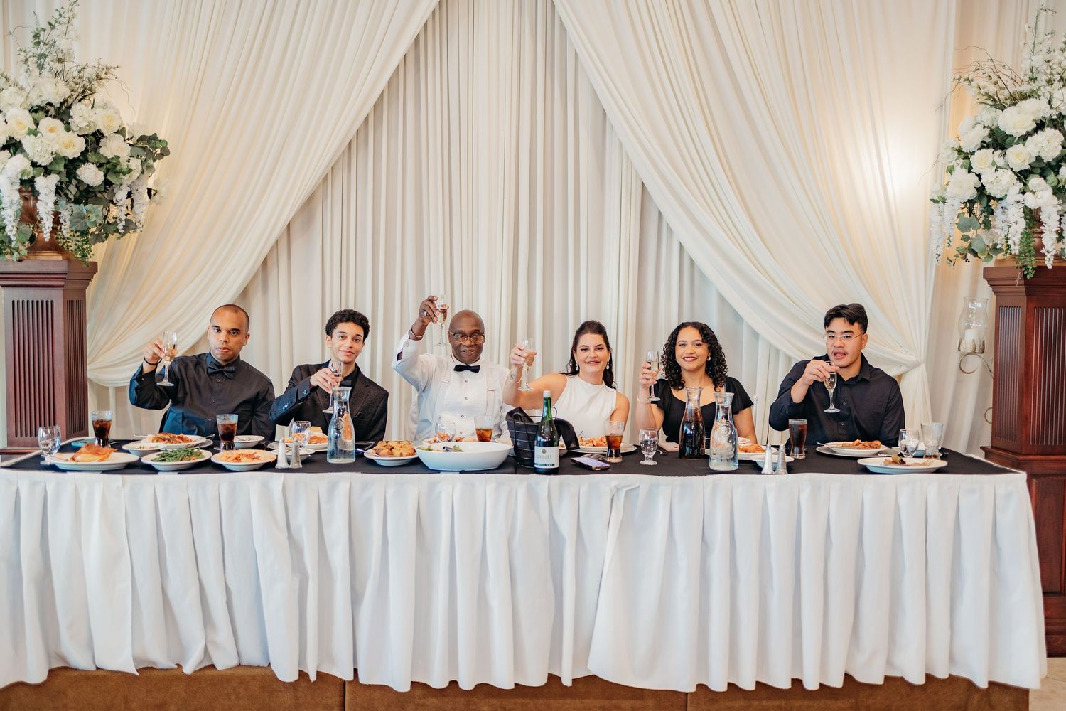 Wedding party members sit at an elegant white head table with floral centerpieces in a reception venue.