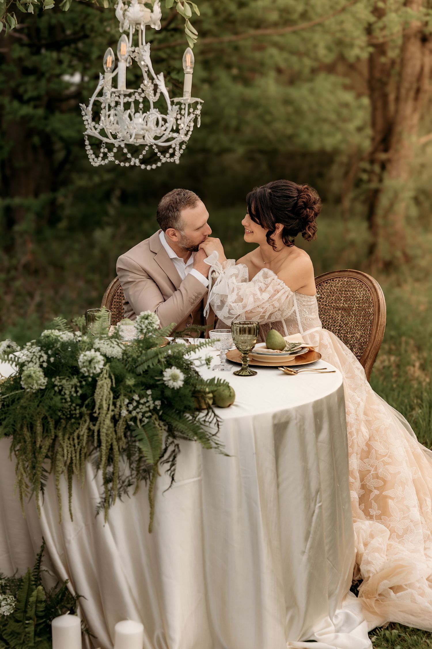 Romantic outdoor table setting with crystal chandelier, white linens, greenery and rattan chair for intimate wedding reception.