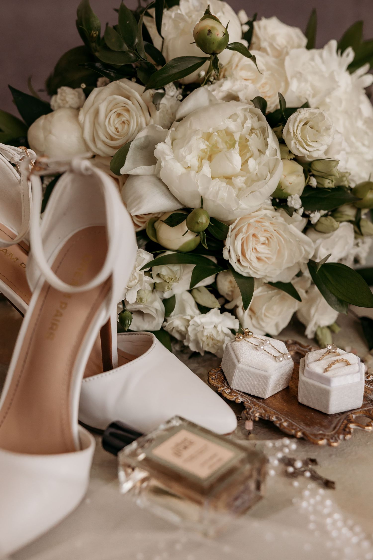 Elegant wedding details featuring white roses, high heel shoes, perfume bottle and small gift boxes on beige surface.