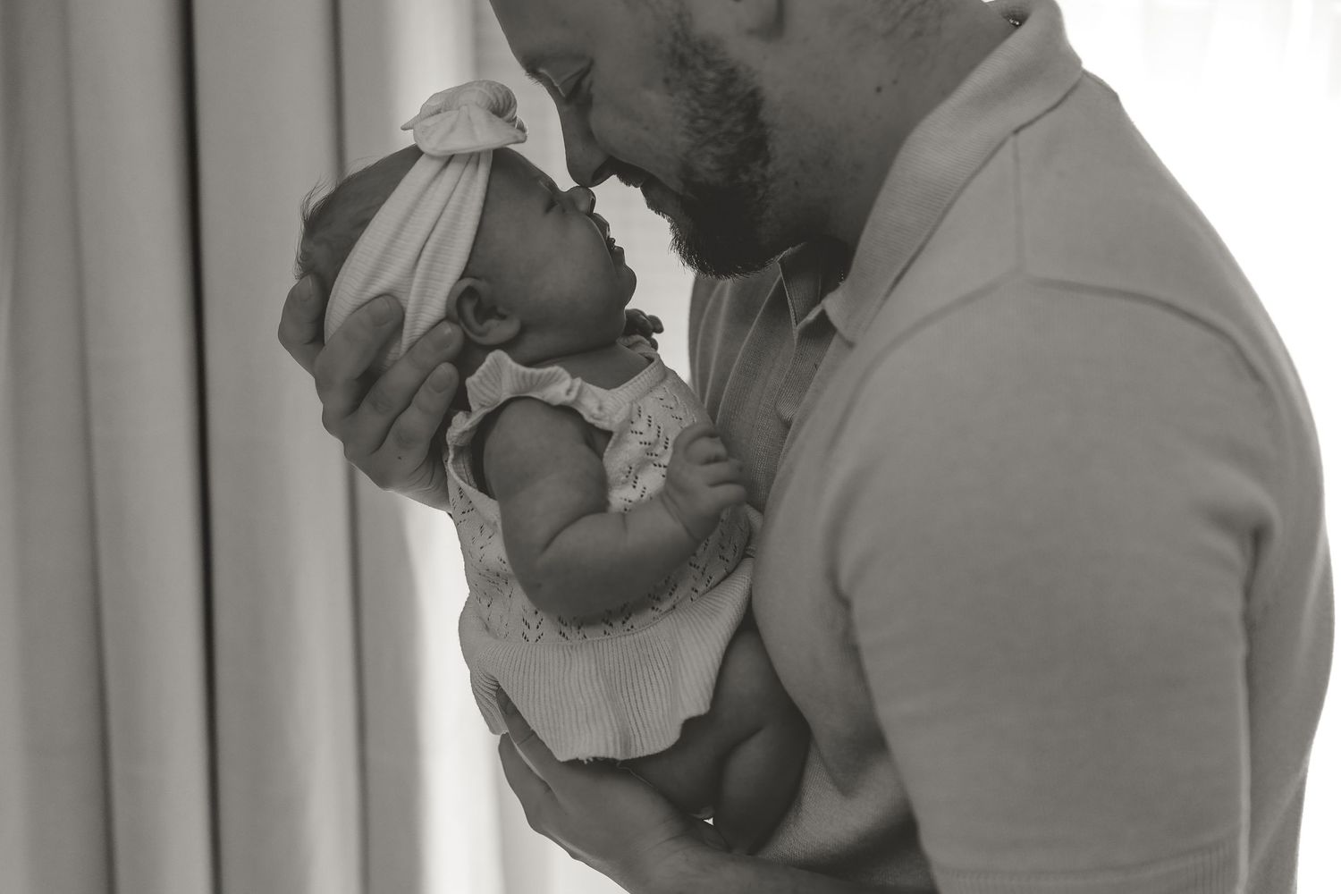 A tender black and white photograph shows a parent in a light polo shirt holding and kissing a tiny newborn baby.