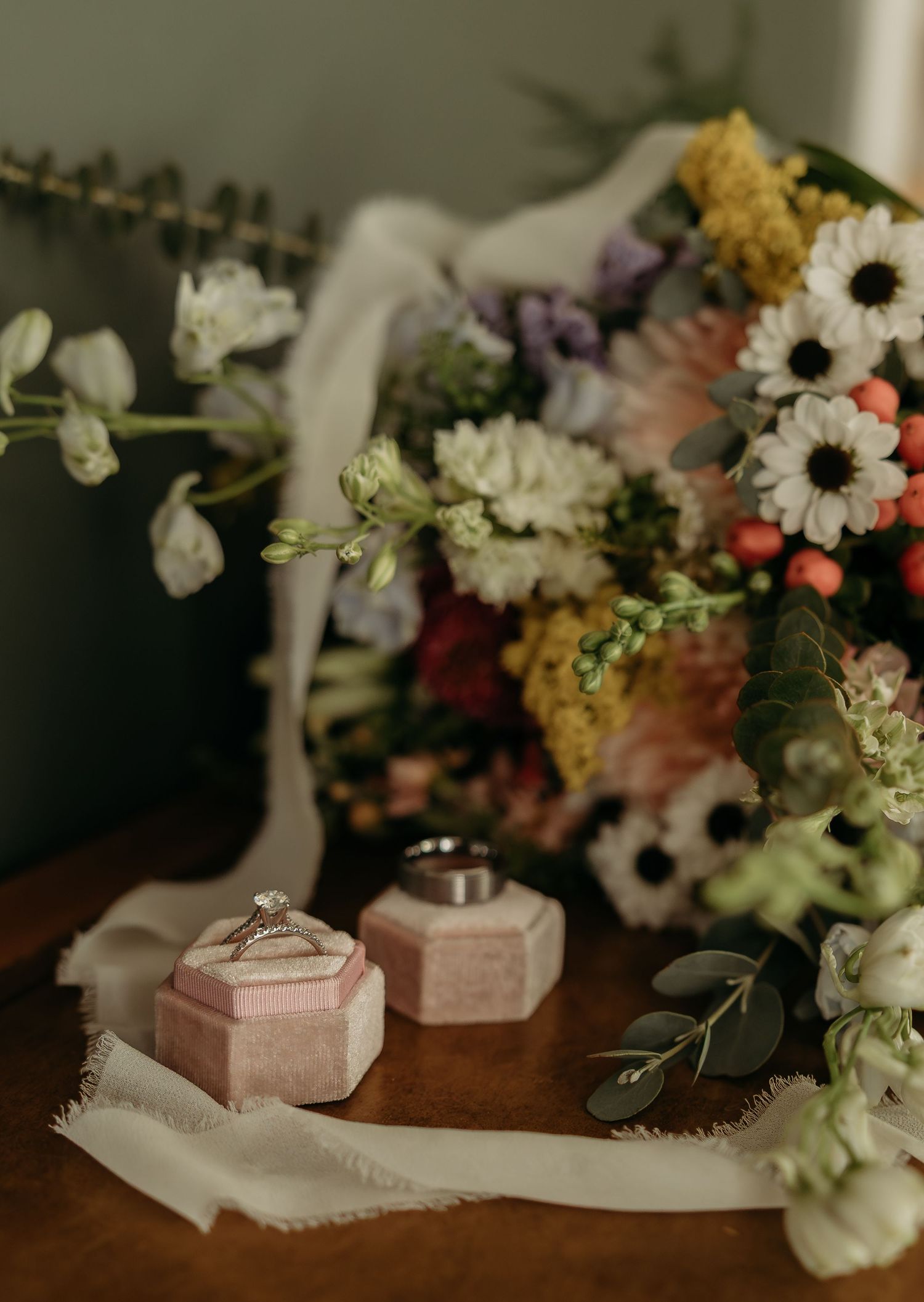 Wedding rings in pink velvet boxes beside a colorful bridal bouquet with white, yellow, and pink flowers on a wooden table.