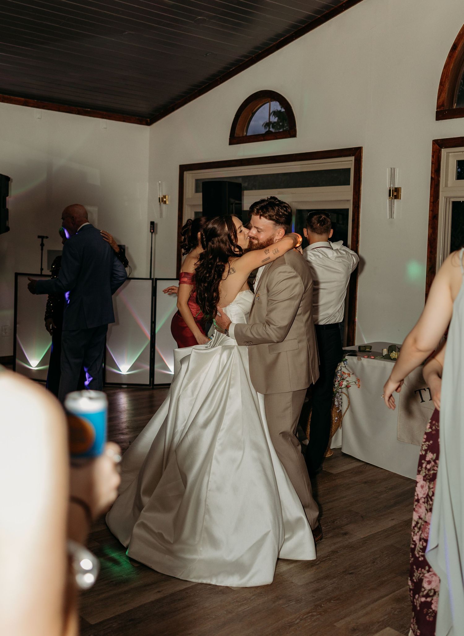 Bride and groom share a romantic first dance kiss at their wedding reception with guests dancing in the background.
