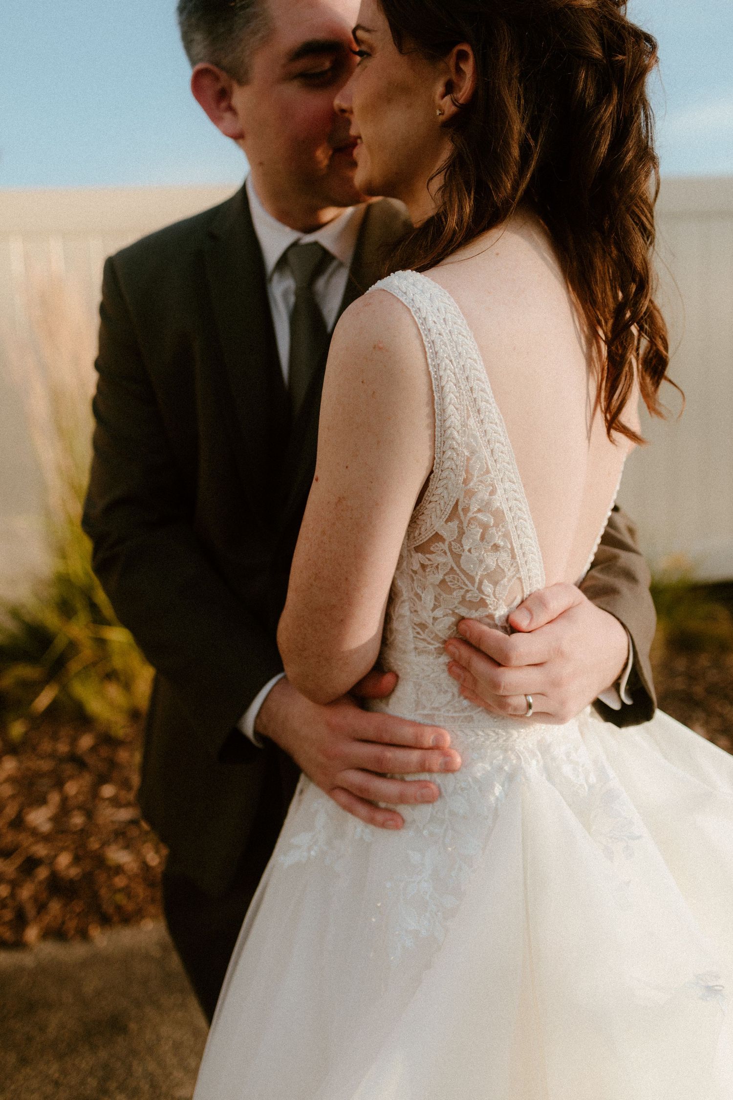 A groom in black suit tenderly holds bride in delicate lace wedding dress during golden hour.